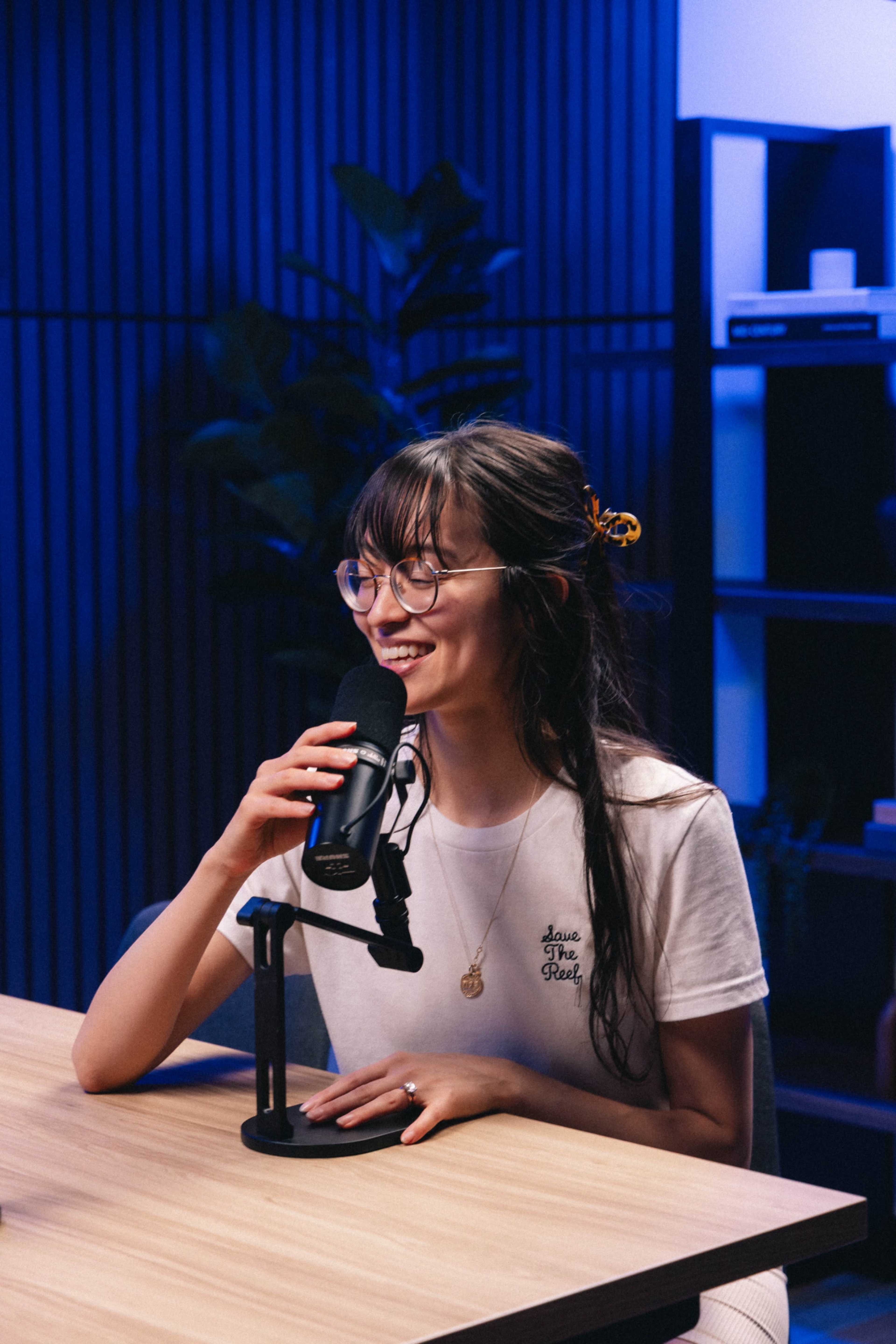A young woman wearing glasses speaks into a microphone while sitting at a wooden table in a modern studio setting.