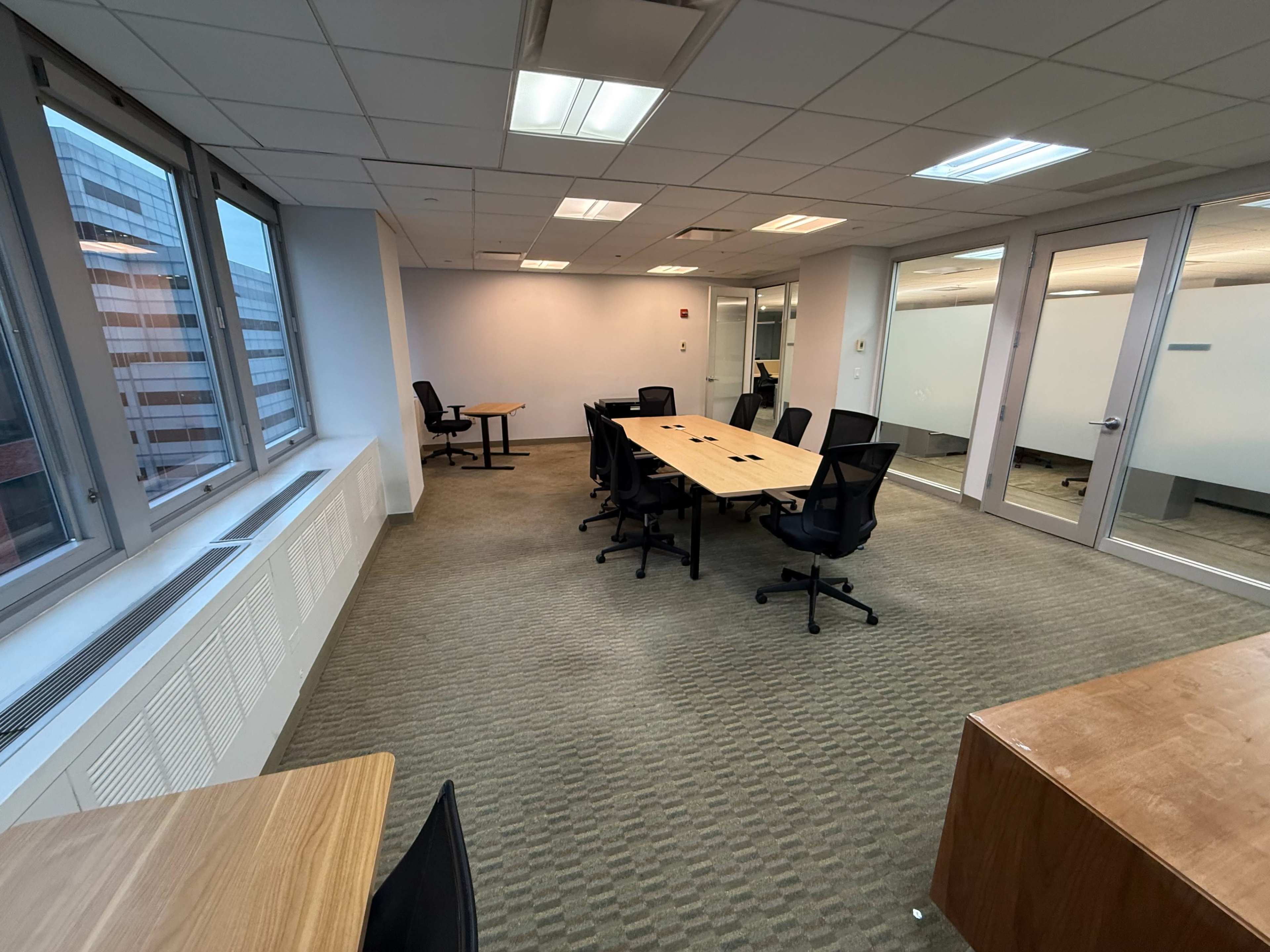 The image shows a modern office space with multiple tables and chairs, large windows, and partitions.