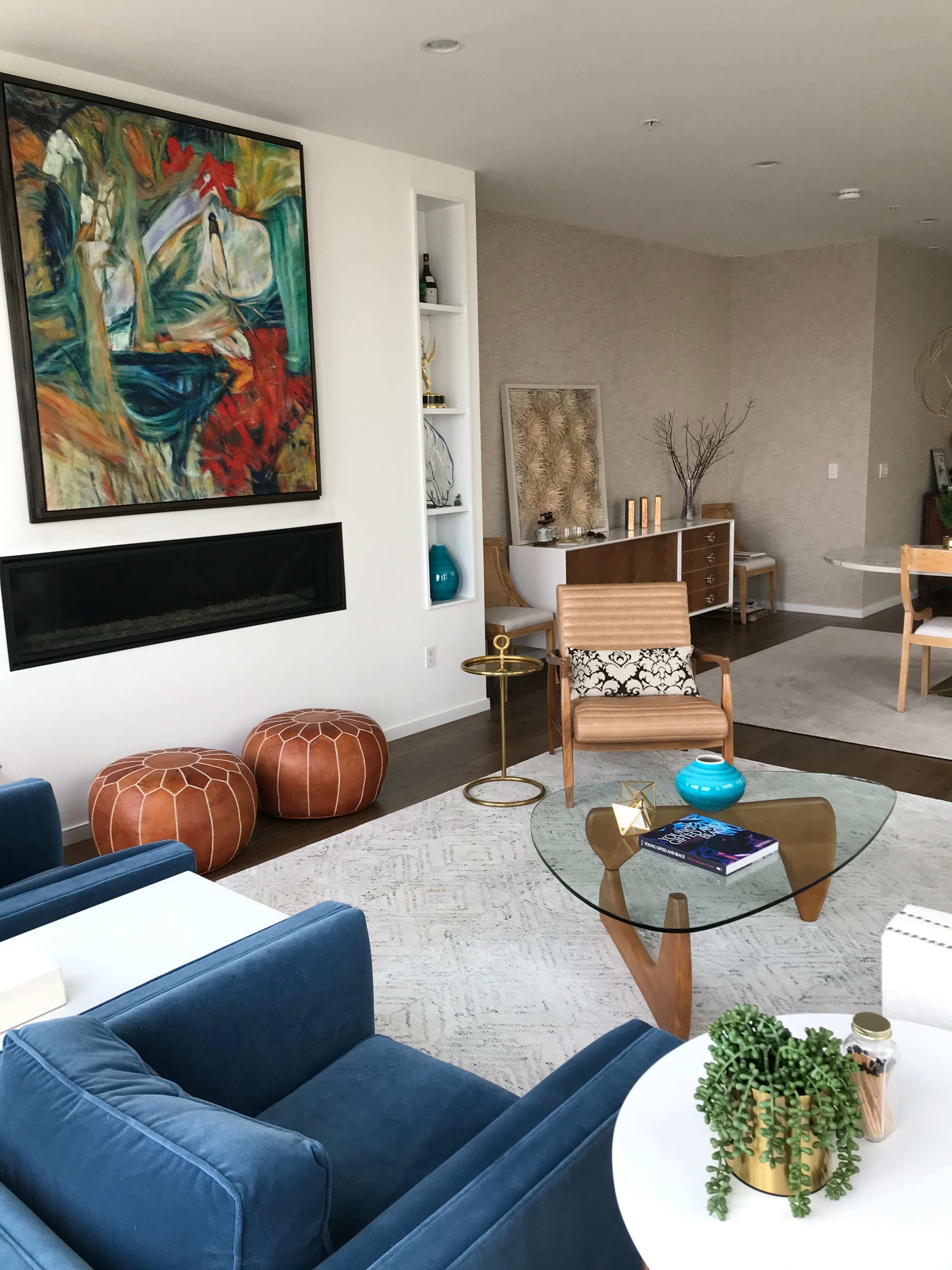 A modern living room features a large abstract painting, a glass coffee table, blue sofas, and decorative elements like poufs and plants.