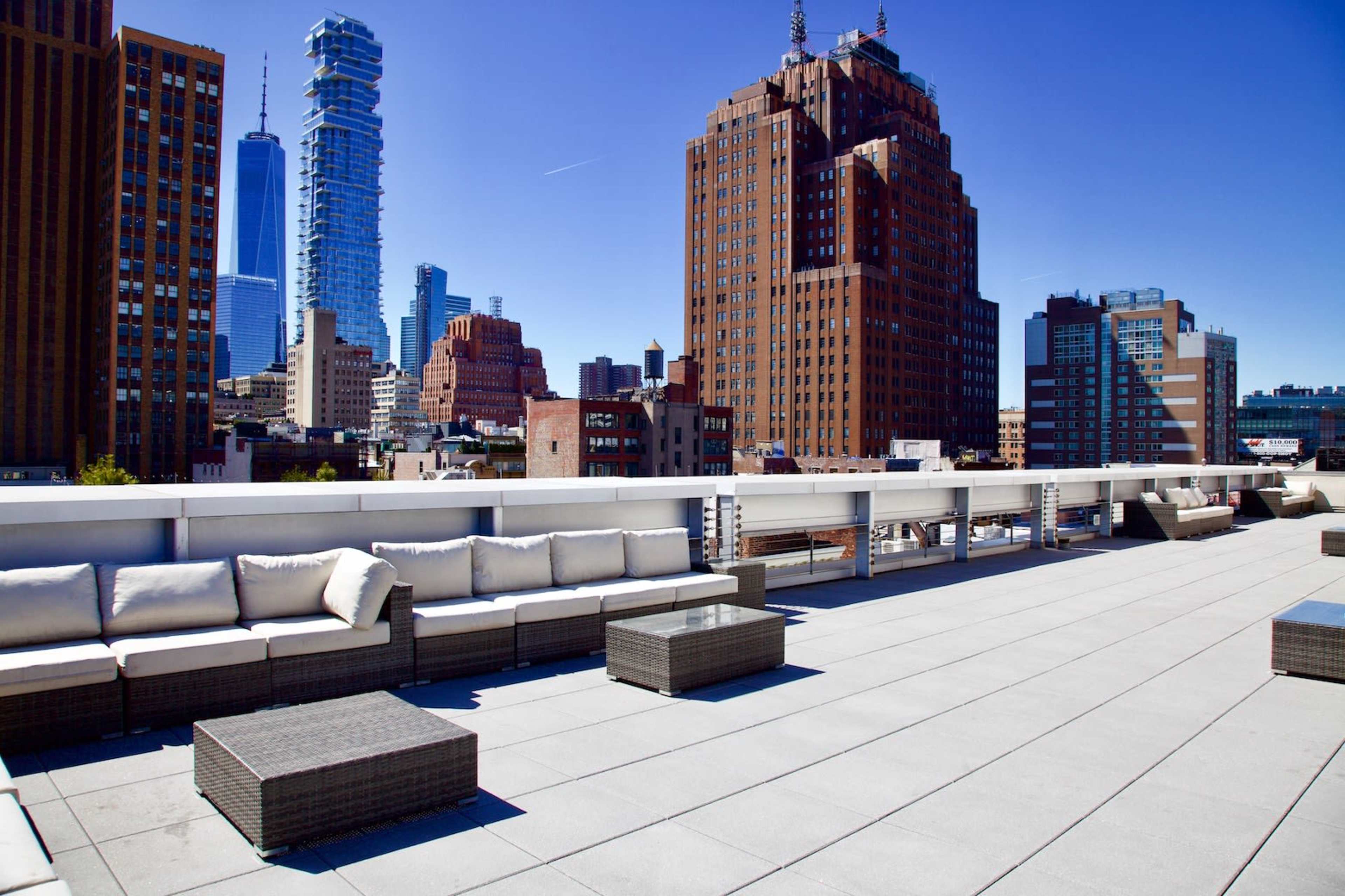 The image shows a rooftop terrace with modern seating and a panoramic view of a city skyline featuring various skyscrapers.