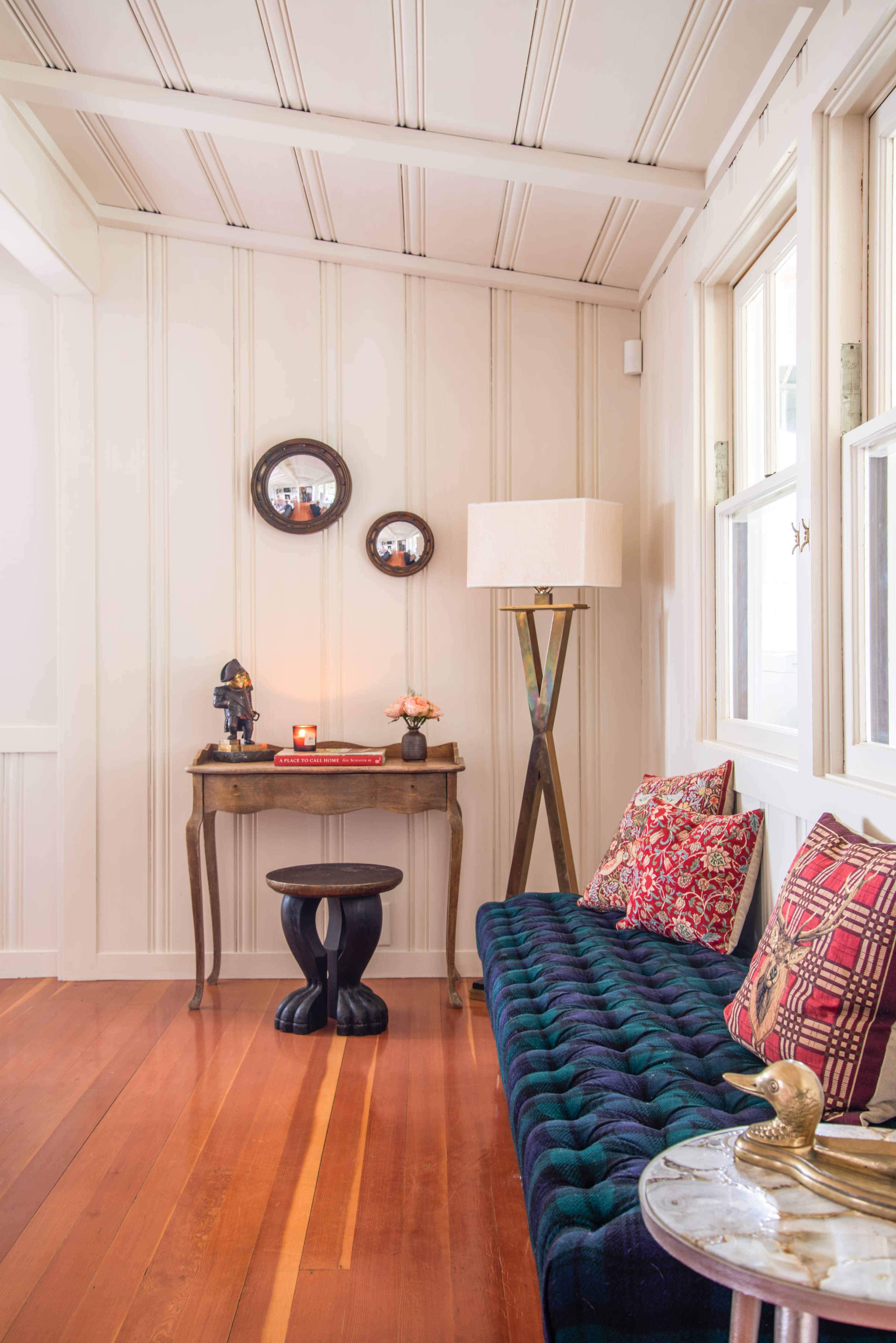 A cozy corner of a room featuring a blue tufted bench, a small wooden table with a lamp, decorative mirrors on the wall, and a polished wooden floor.