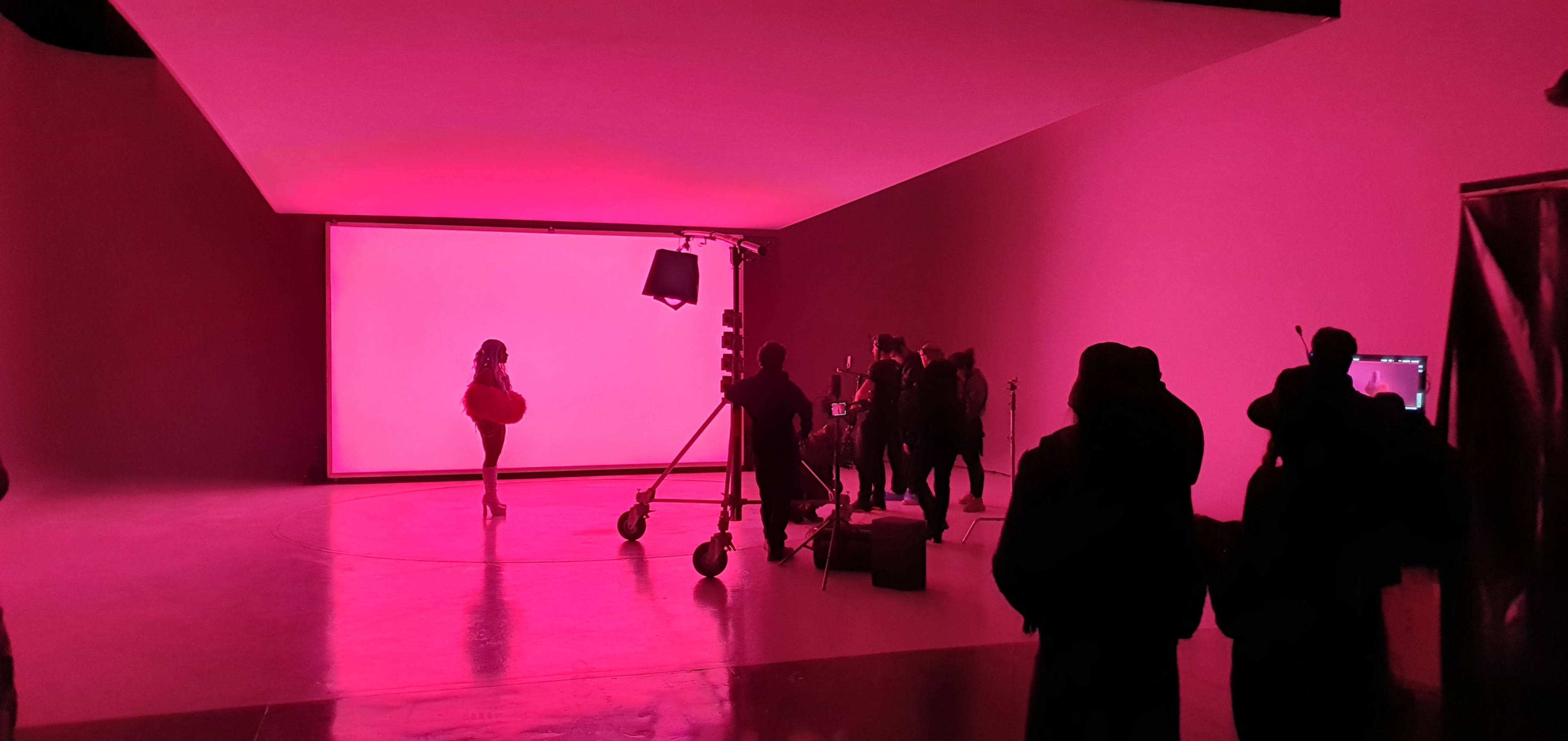 A group of people is gathered in a studio with pink lighting, while a model poses in front of a white backdrop.