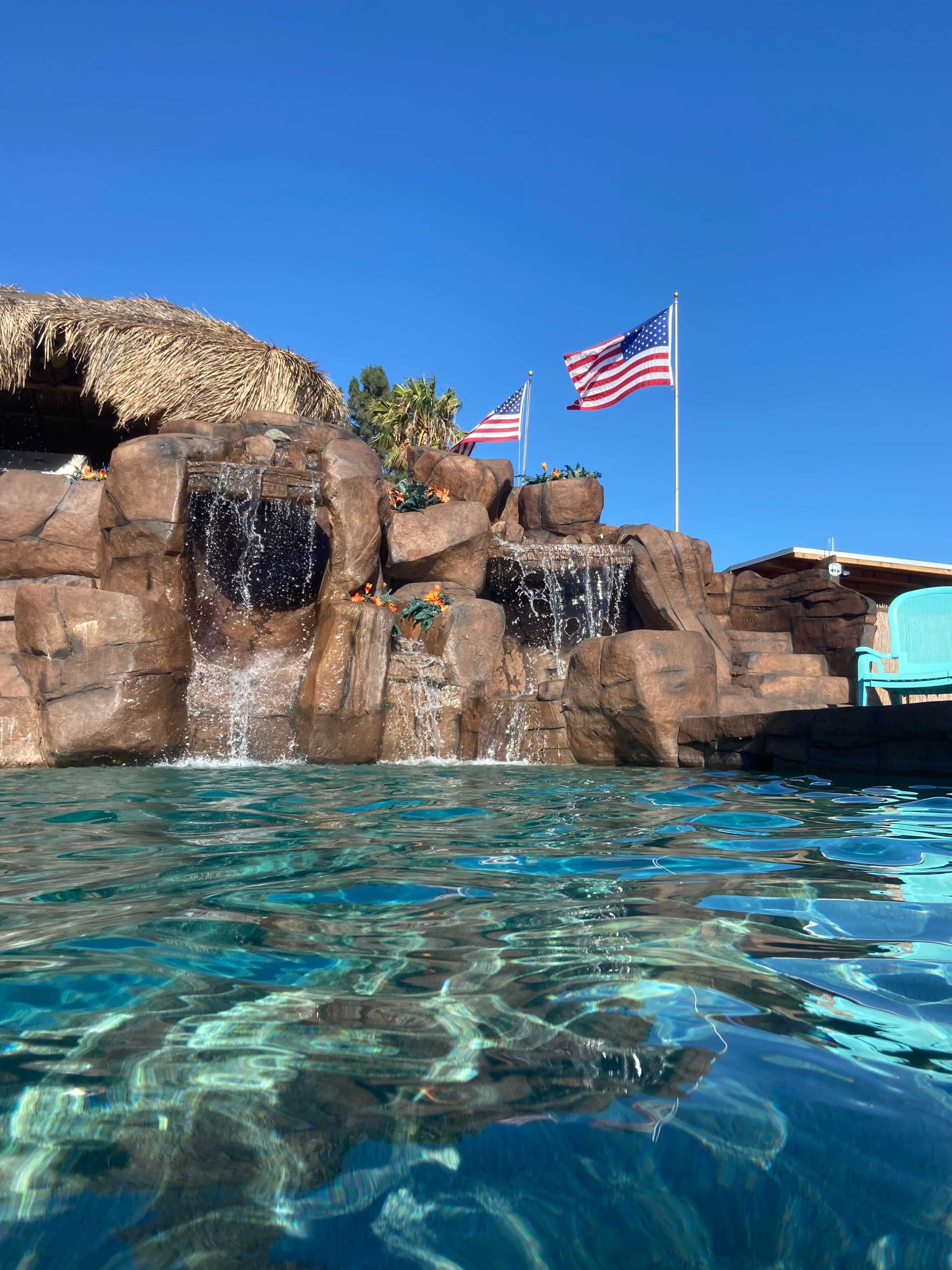 A swimming pool with a rock waterfall and two American flags waving in the background against a clear blue sky.