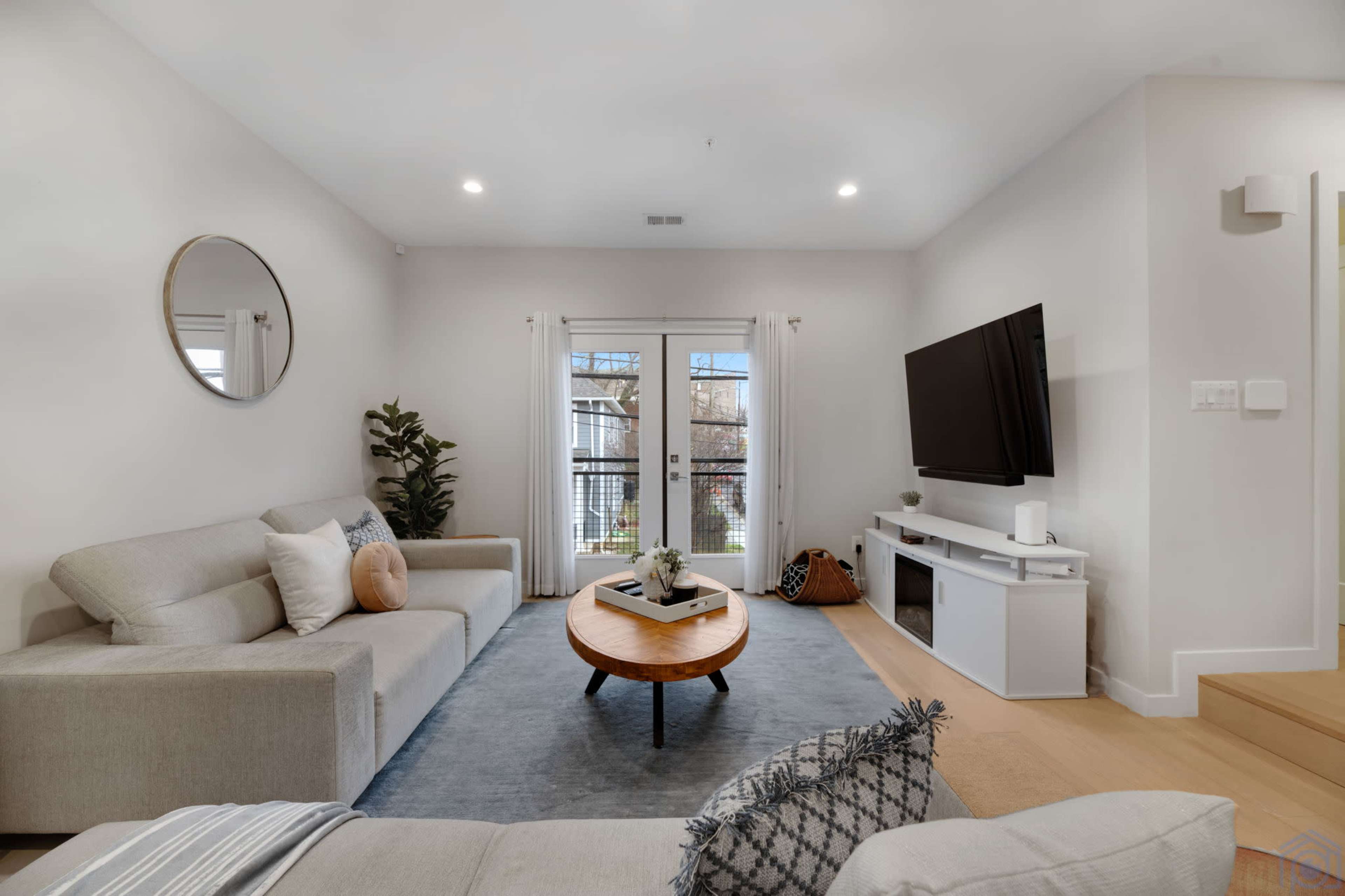 A modern living room features a light gray sectional sofa, a round coffee table, and a wall-mounted television, with large windows overlooking a balcony.