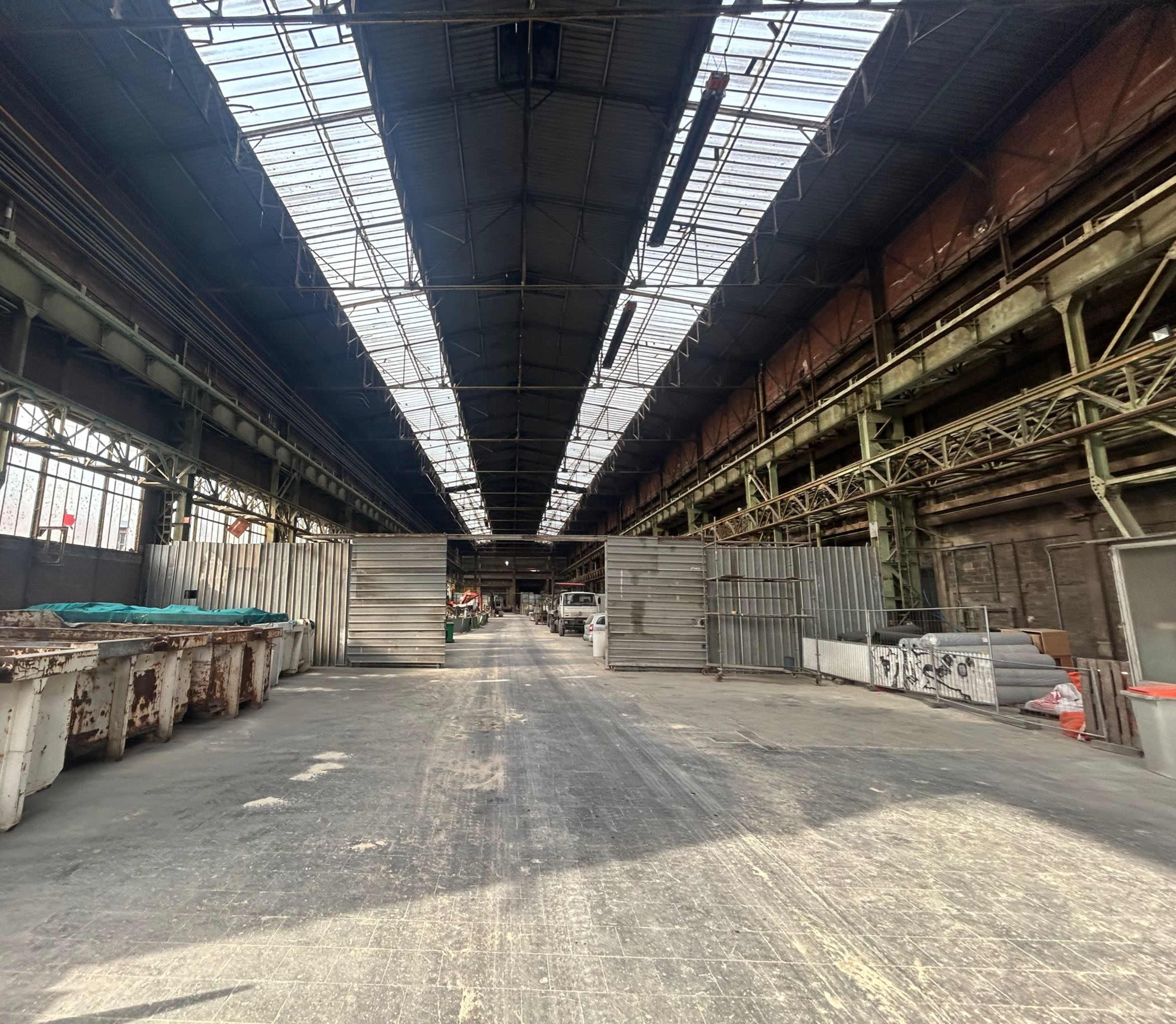 A long, industrial warehouse interior with a clear path running down the center, flanked by metal containers and large overhead windows allowing natural light to enter.
