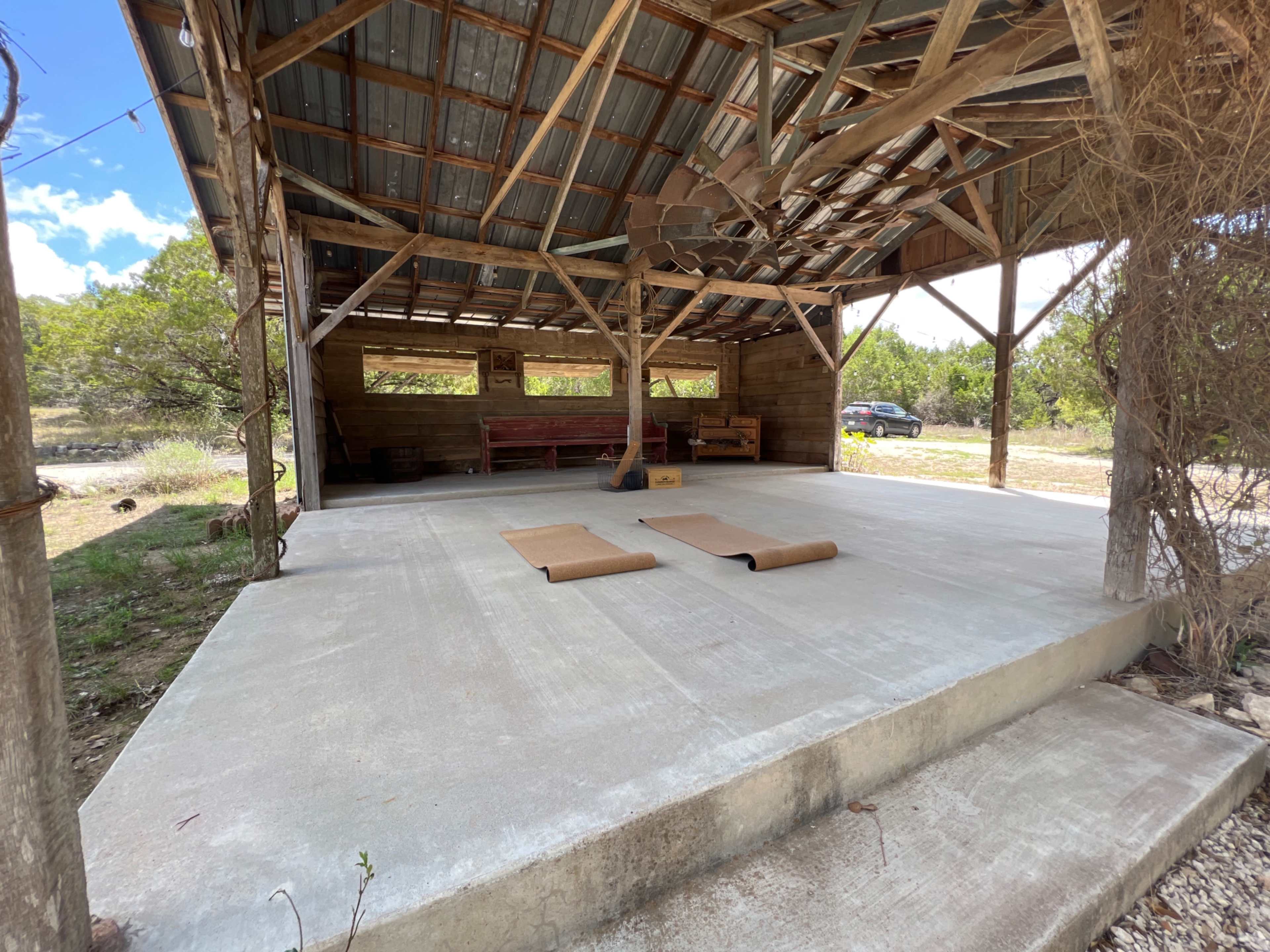 A covered outdoor stage features two yoga mats laid out on a concrete floor, surrounded by greenery and a parked car visible in the background.