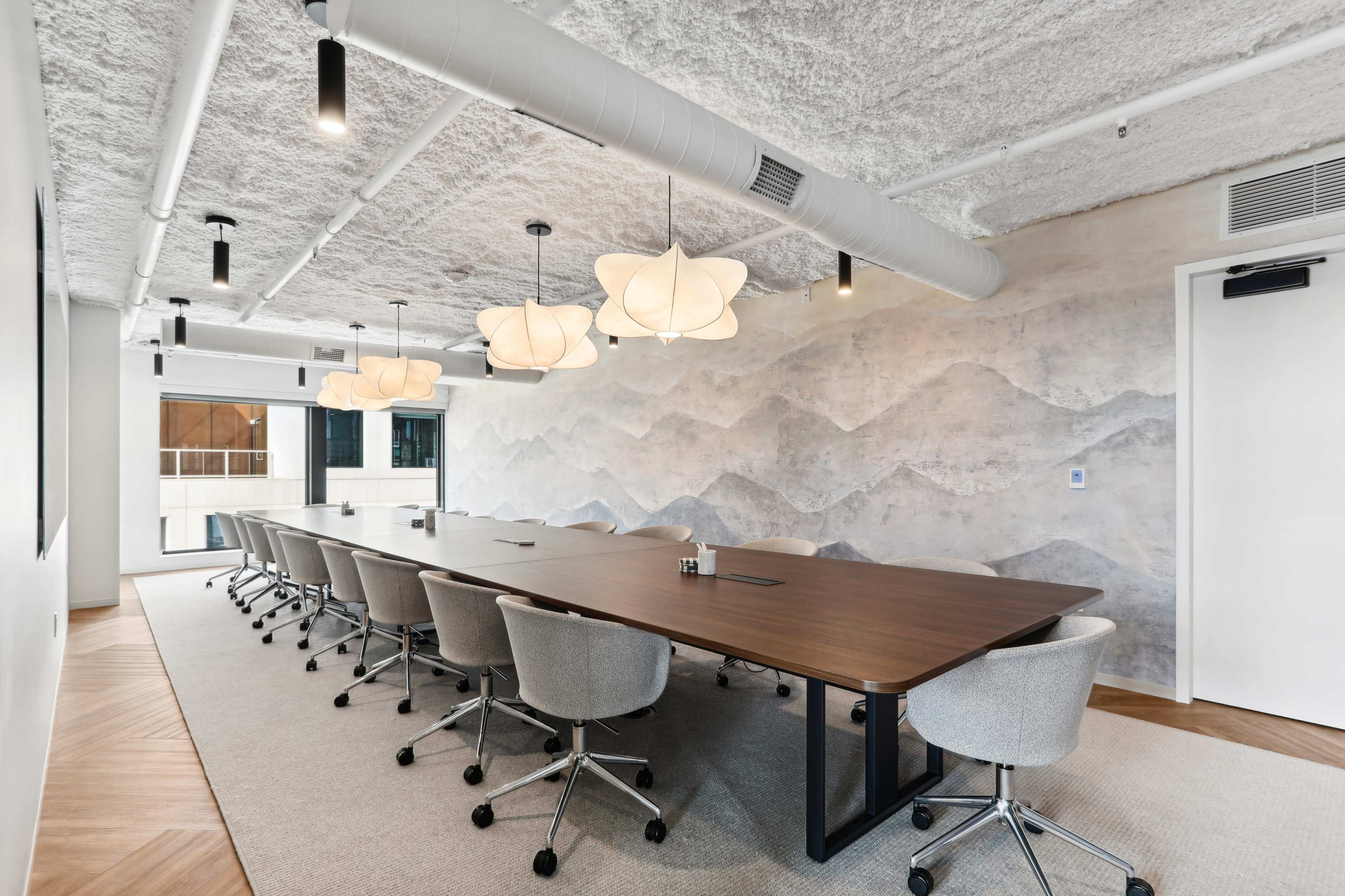 A modern conference room features a long wooden table surrounded by ergonomic chairs, with soft lighting and a textured wall depicting a mountain range.