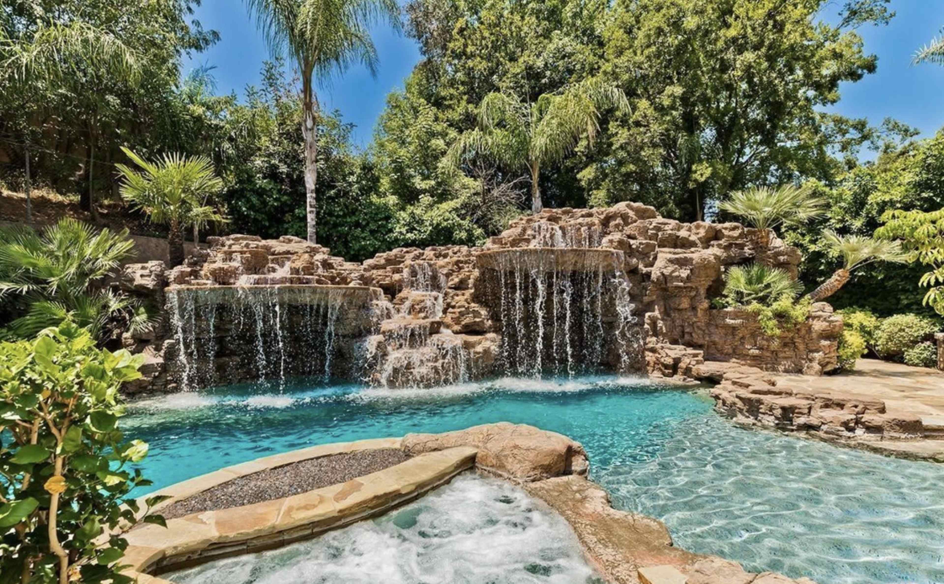 The image shows a landscaped pool area featuring cascading waterfalls and a rocky formation surrounded by lush greenery.