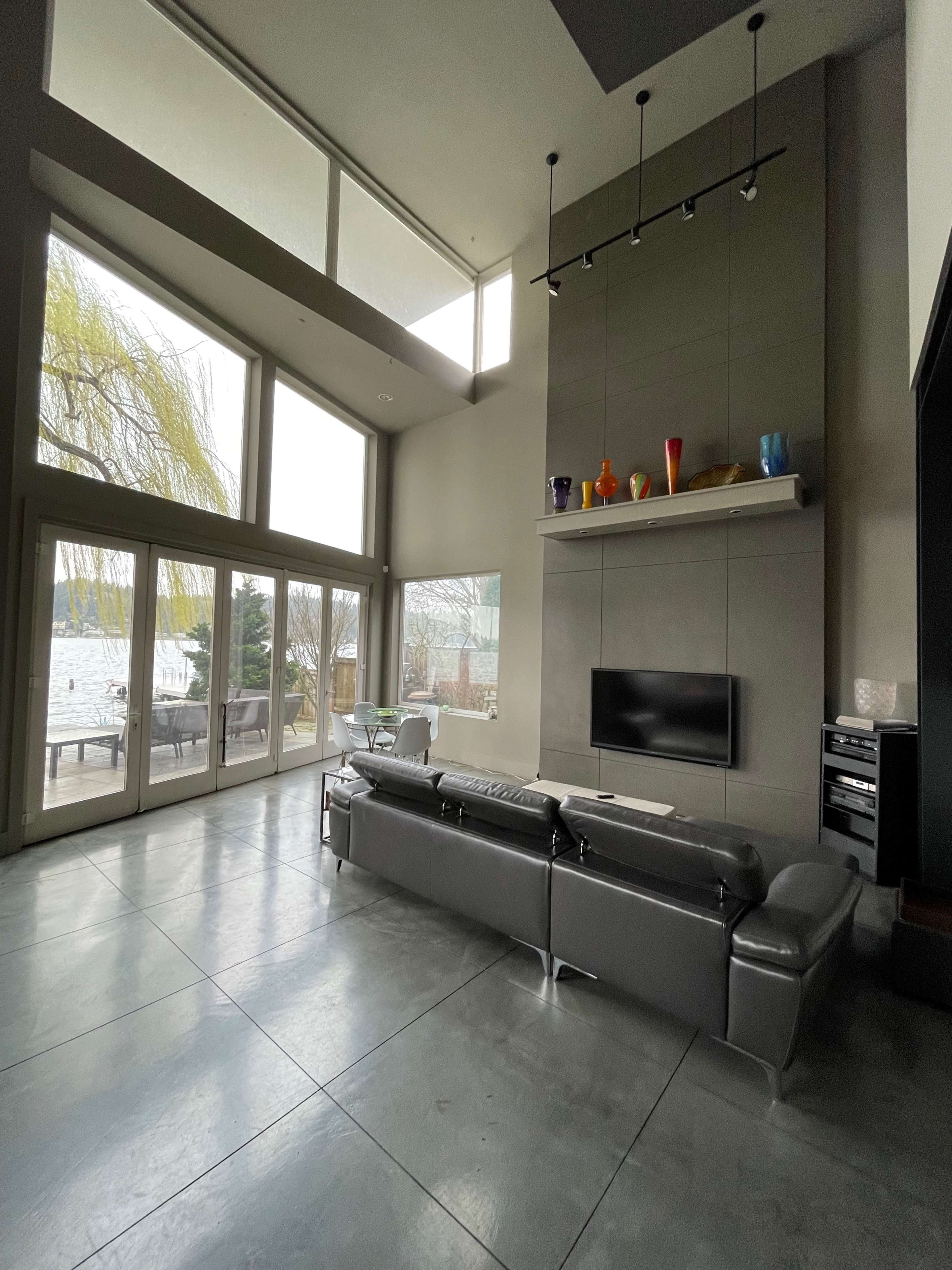A modern living room with a large sectional sofa, a wall-mounted television, and large windows overlooking a body of water.