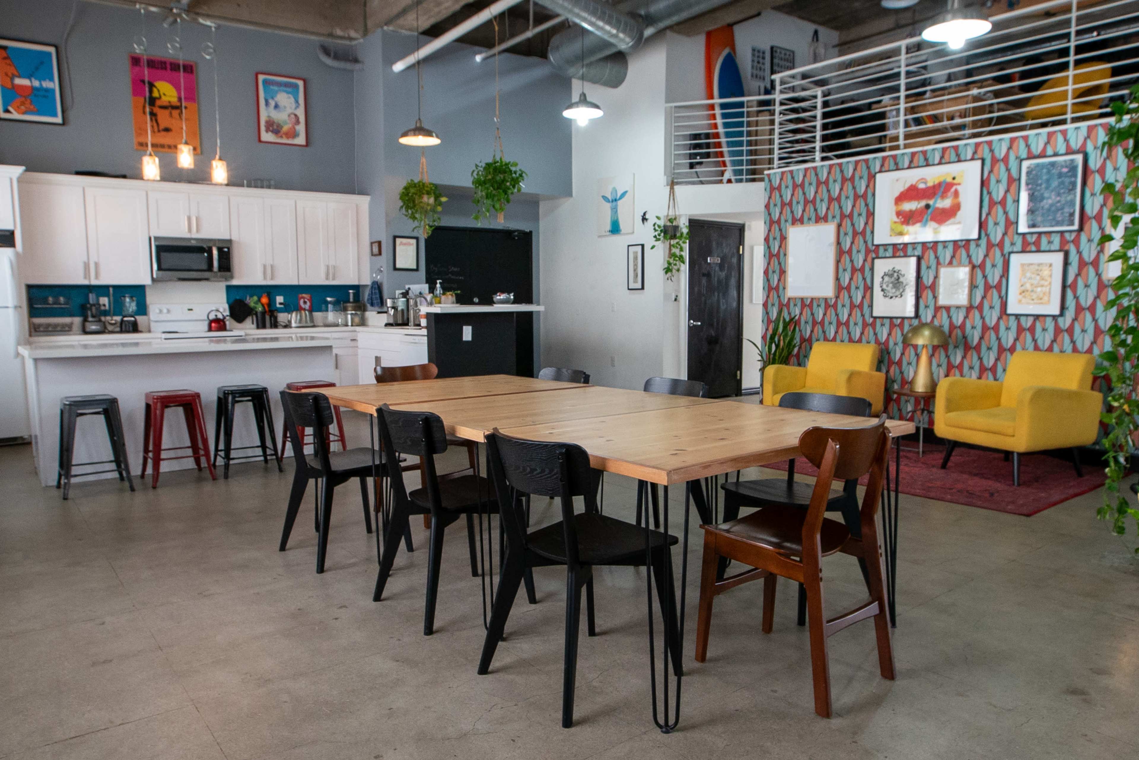 A spacious communal area with a long wooden table surrounded by chairs, a kitchenette in the background, and a colorful patterned wall adorned with artwork and plants.