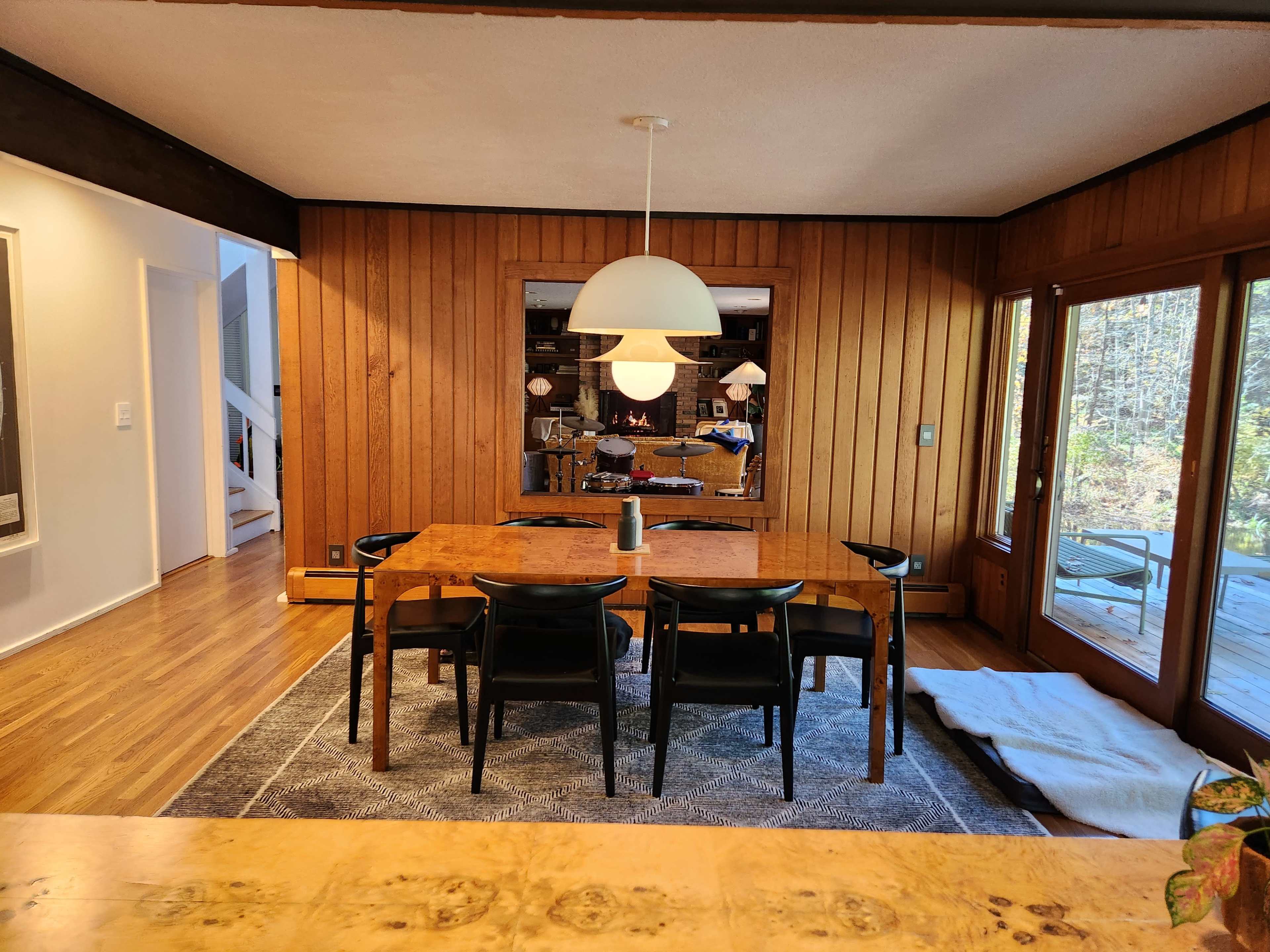 A dining room features a wooden table surrounded by black chairs, with a large pendant light above and wooden wall panels.