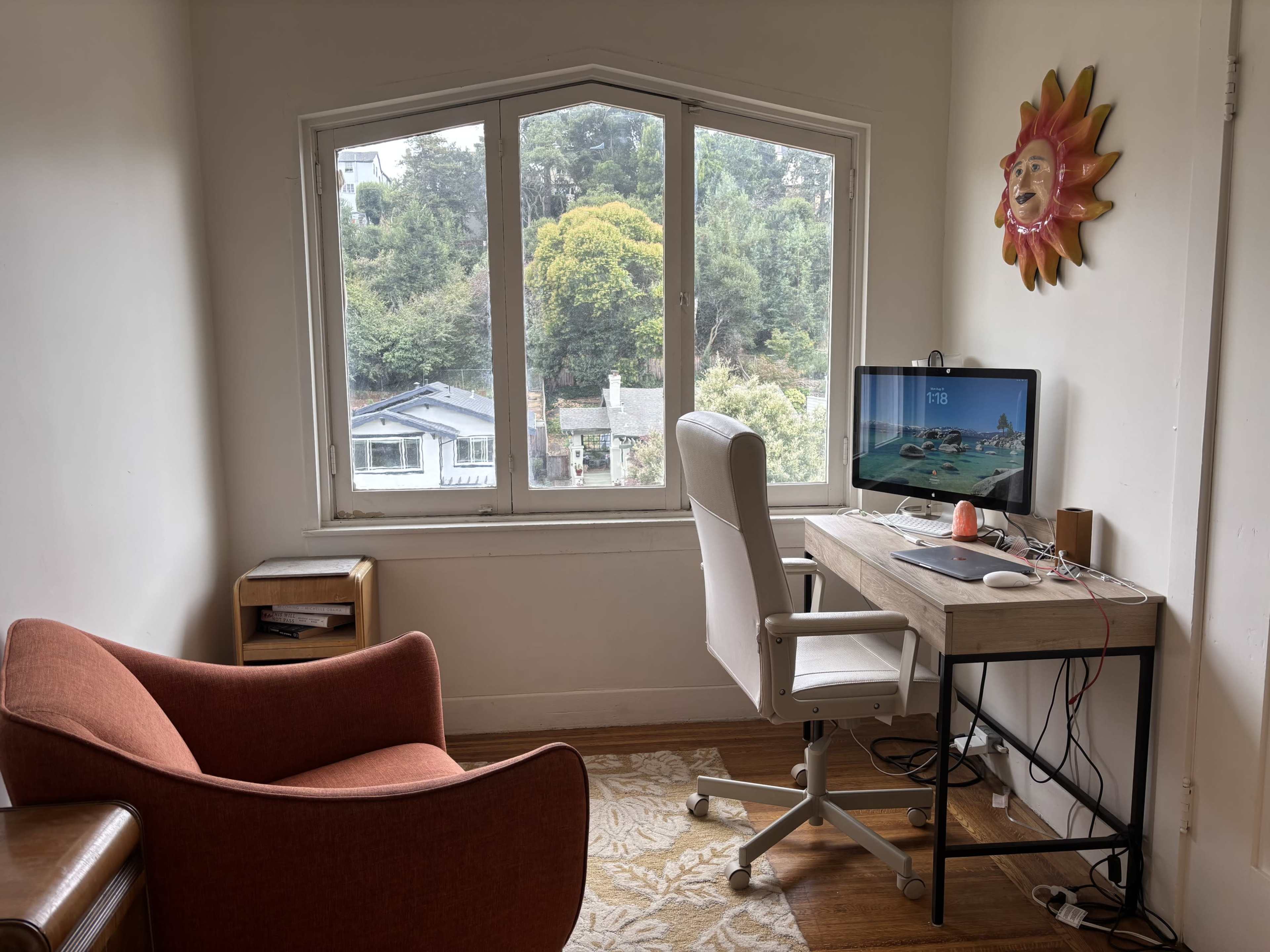 The image shows a small home office with a desk and computer set against a large window overlooking trees and a house in the distance.