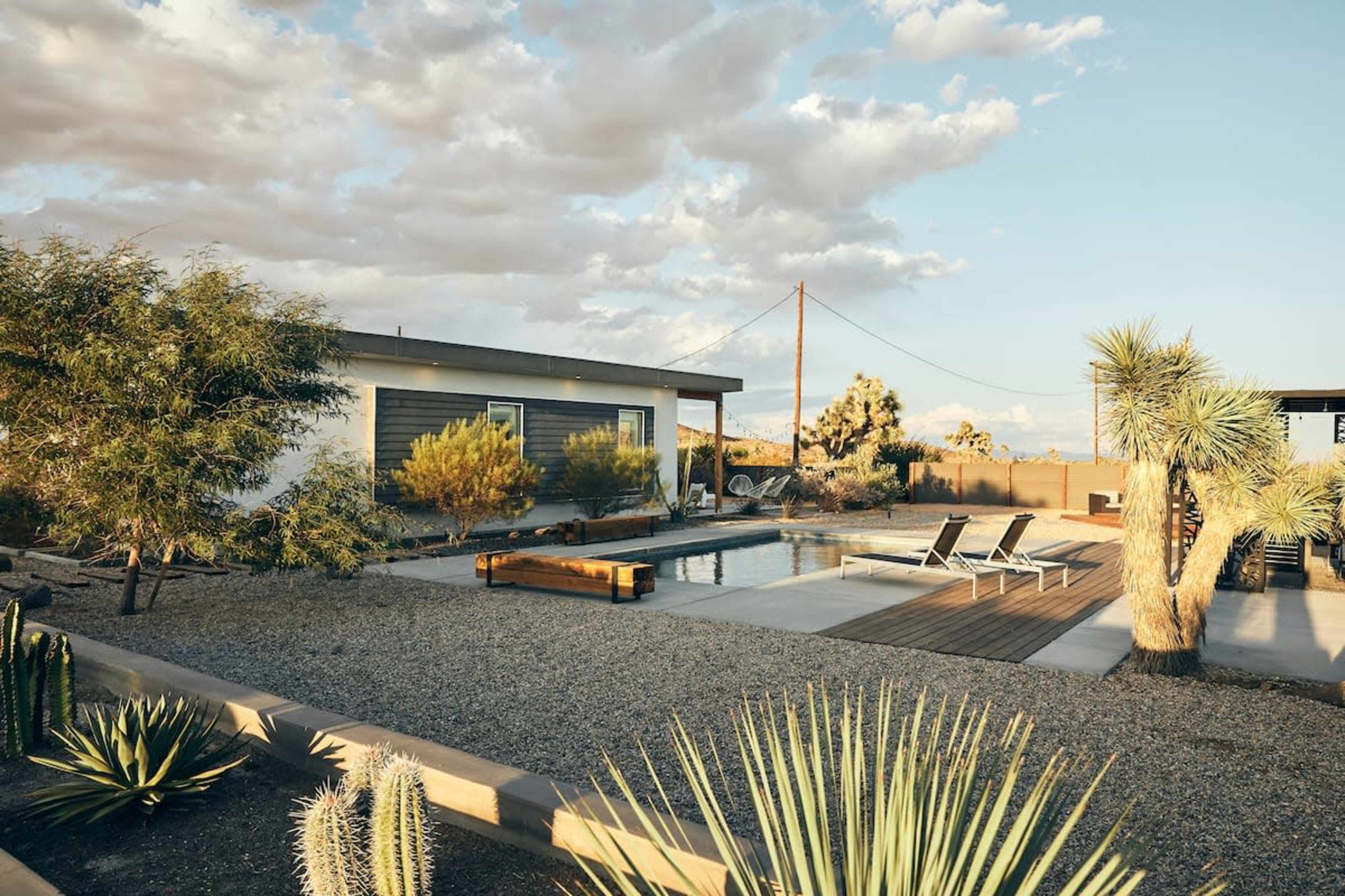 A modern house with a swimming pool is surrounded by gravel landscaping and desert plants under a partly cloudy sky.
