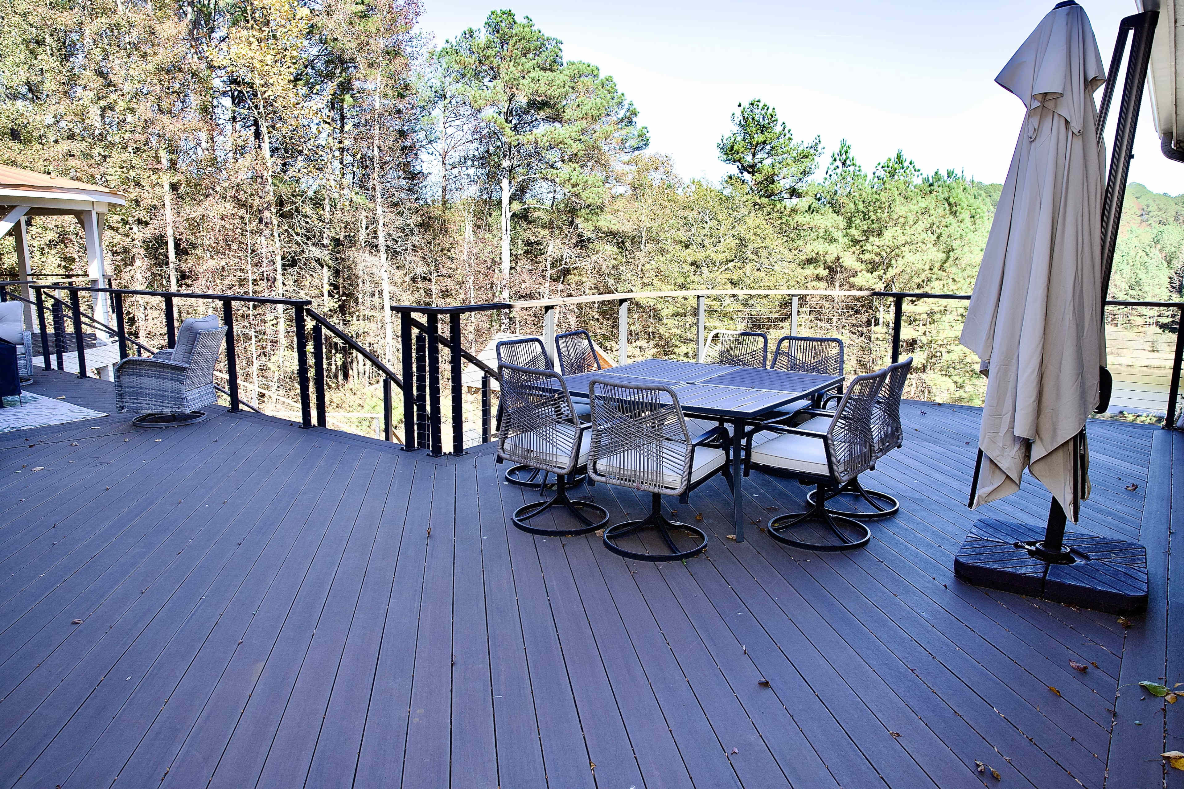 A wooden deck features a table with six chairs, a large umbrella, and a view of surrounding trees.