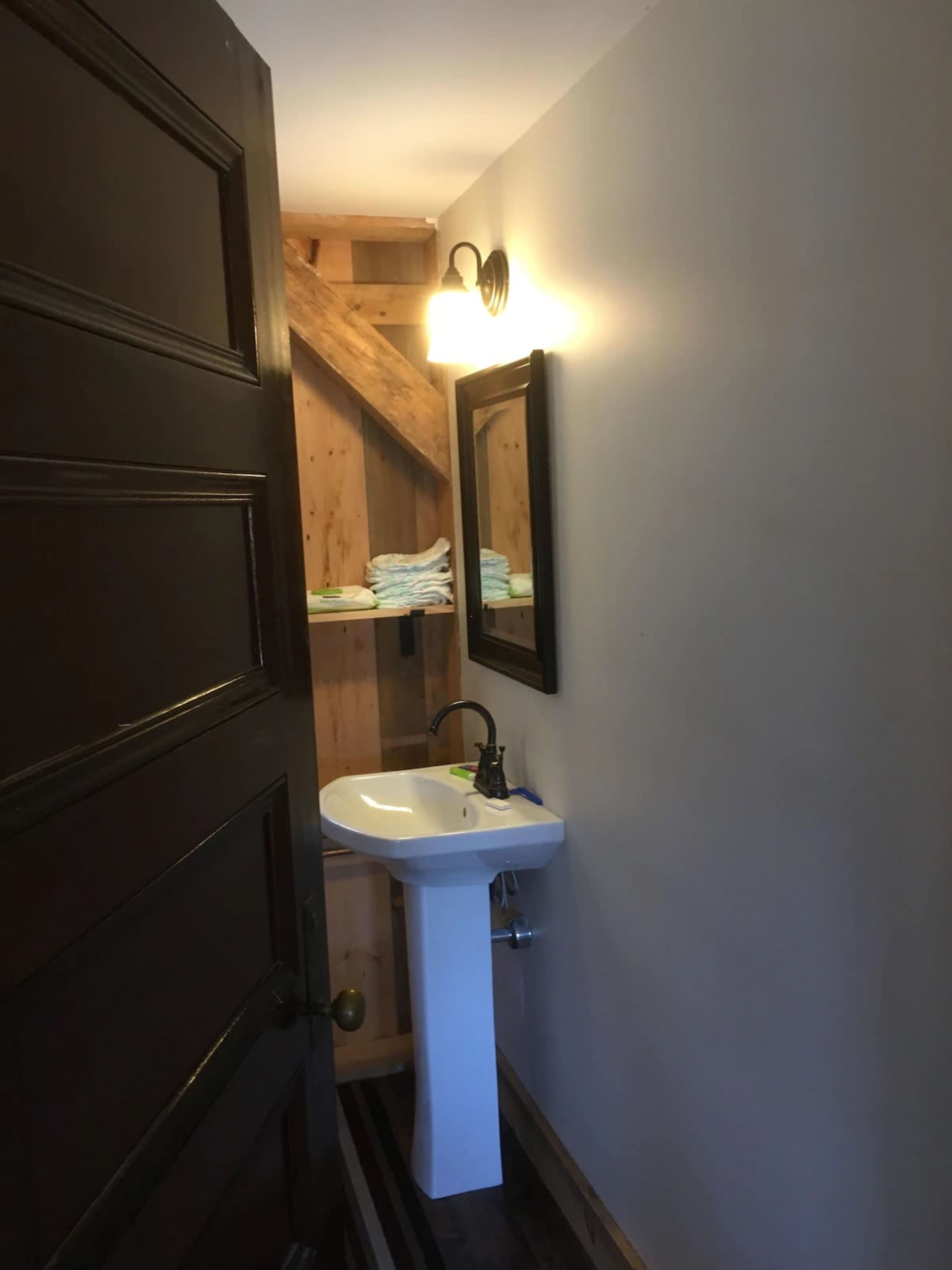 The image shows a small bathroom with a pedestal sink, a mirror, and neatly stacked towels on a shelf, illuminated by a wall-mounted light fixture.