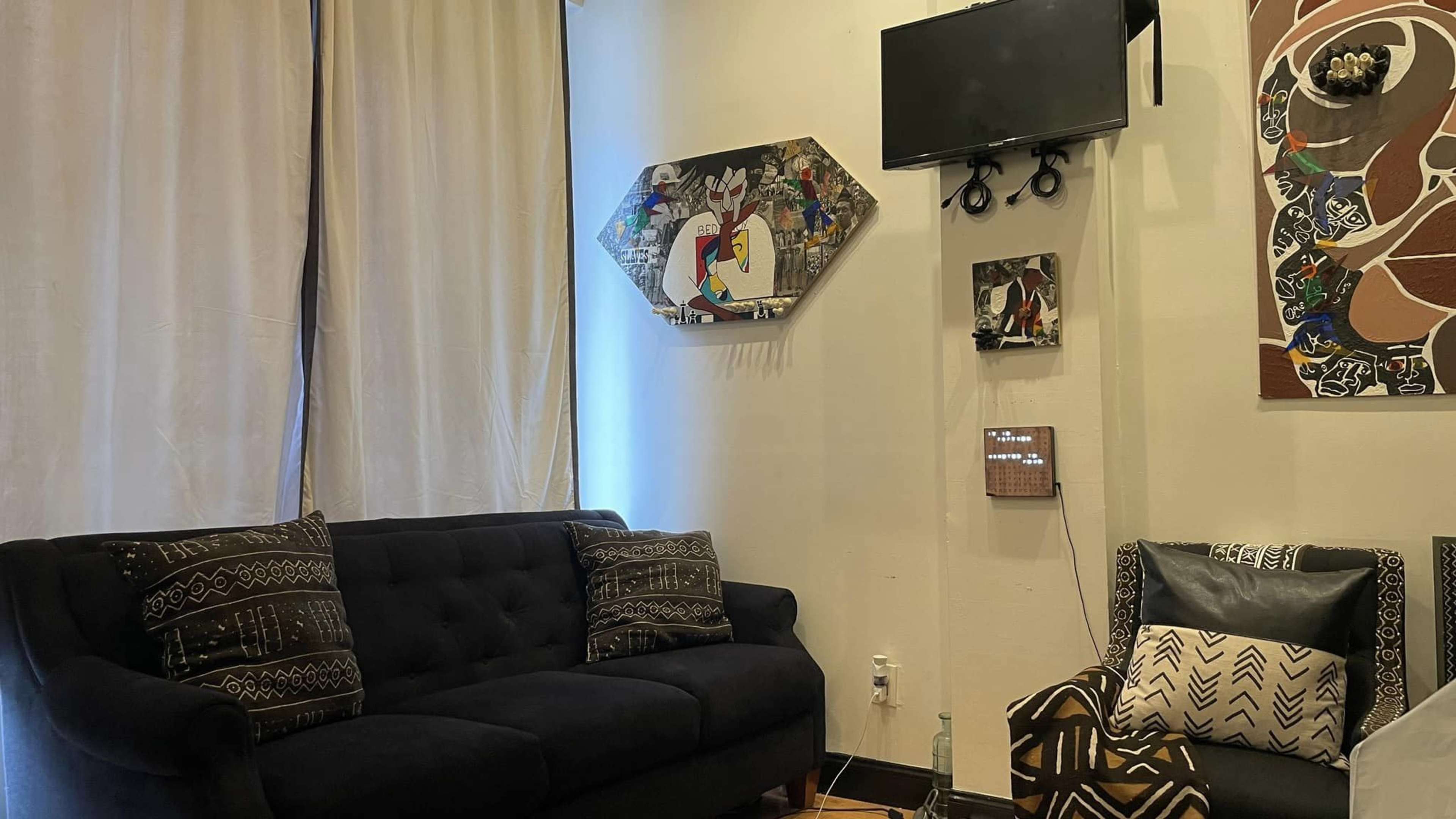 A living room with a dark sofa, patterned throw pillows, a wall-mounted television, and various artworks displayed on the walls.