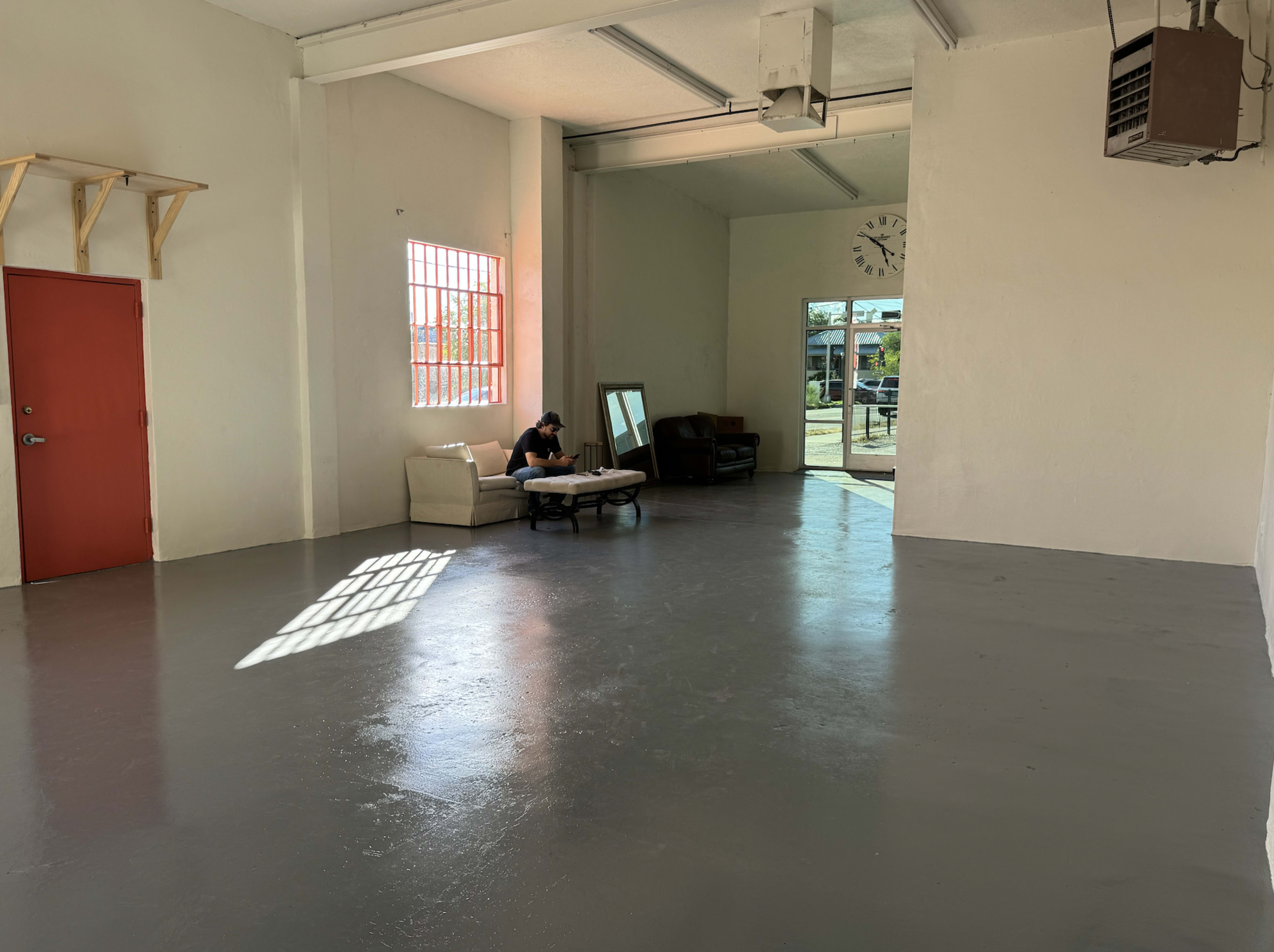 A person sits on a couch in an empty, well-lit room with grey floors and large windows.