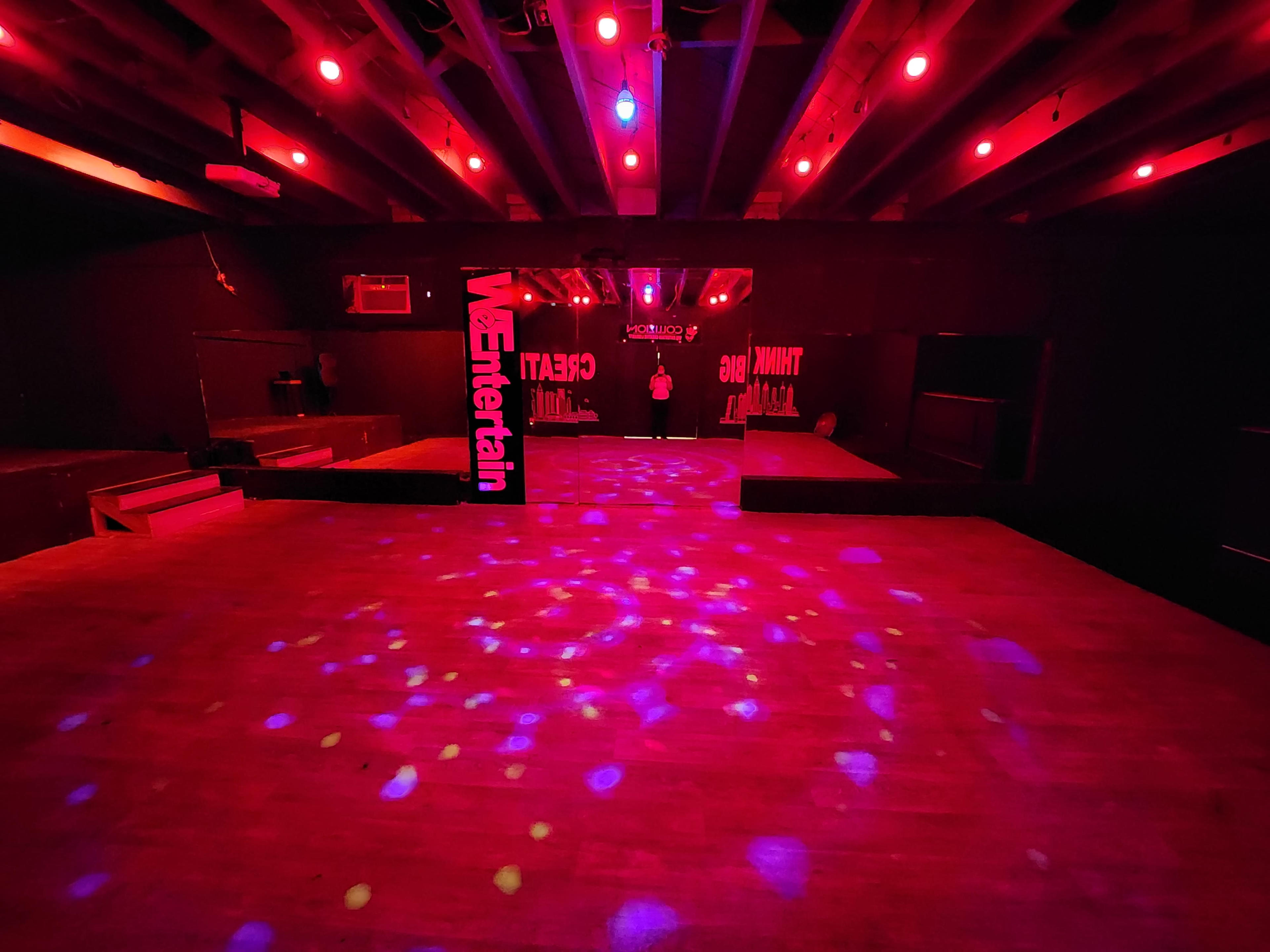 An empty nightclub interior illuminated by red and purple lights, featuring a wooden floor and a sign that reads "We Entertain."