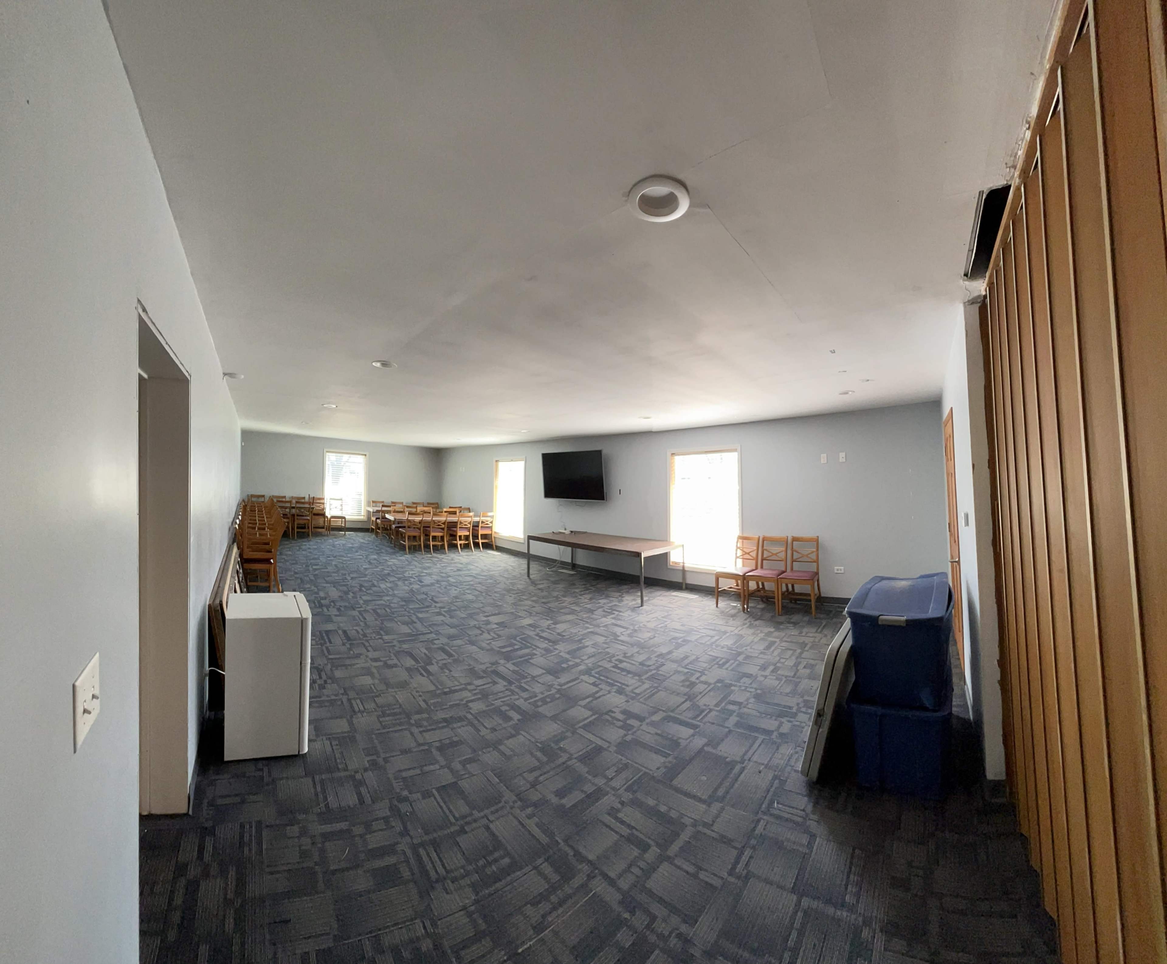 A spacious room with gray walls and a carpeted floor, featuring a table, chairs, a television on the wall, and a few large windows on one side.