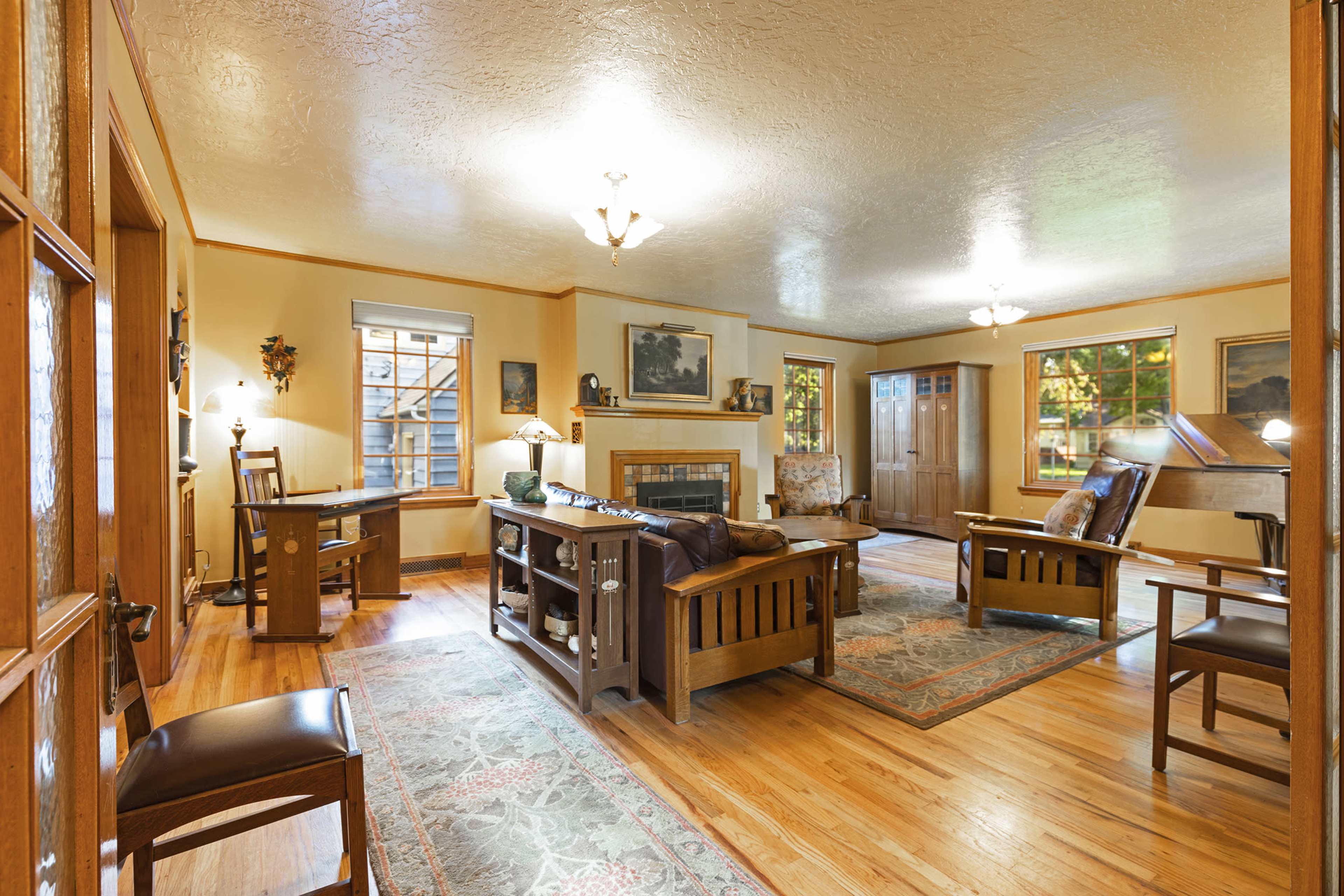 The room features a central seating area with two armchairs, a wooden coffee table, and a fireplace, surrounded by large windows and framed artwork on the walls.