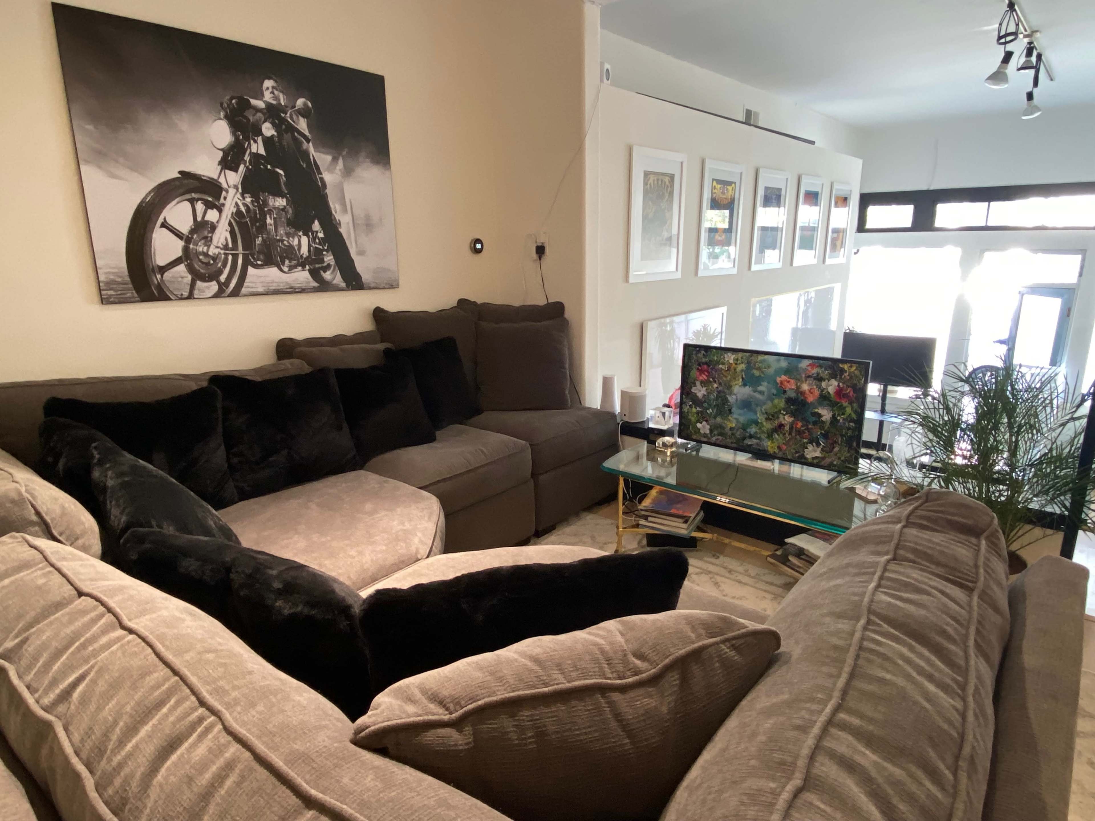 The image shows a living room with a large, sectional couch facing a glass coffee table and a television, alongside a wall adorned with framed artwork.