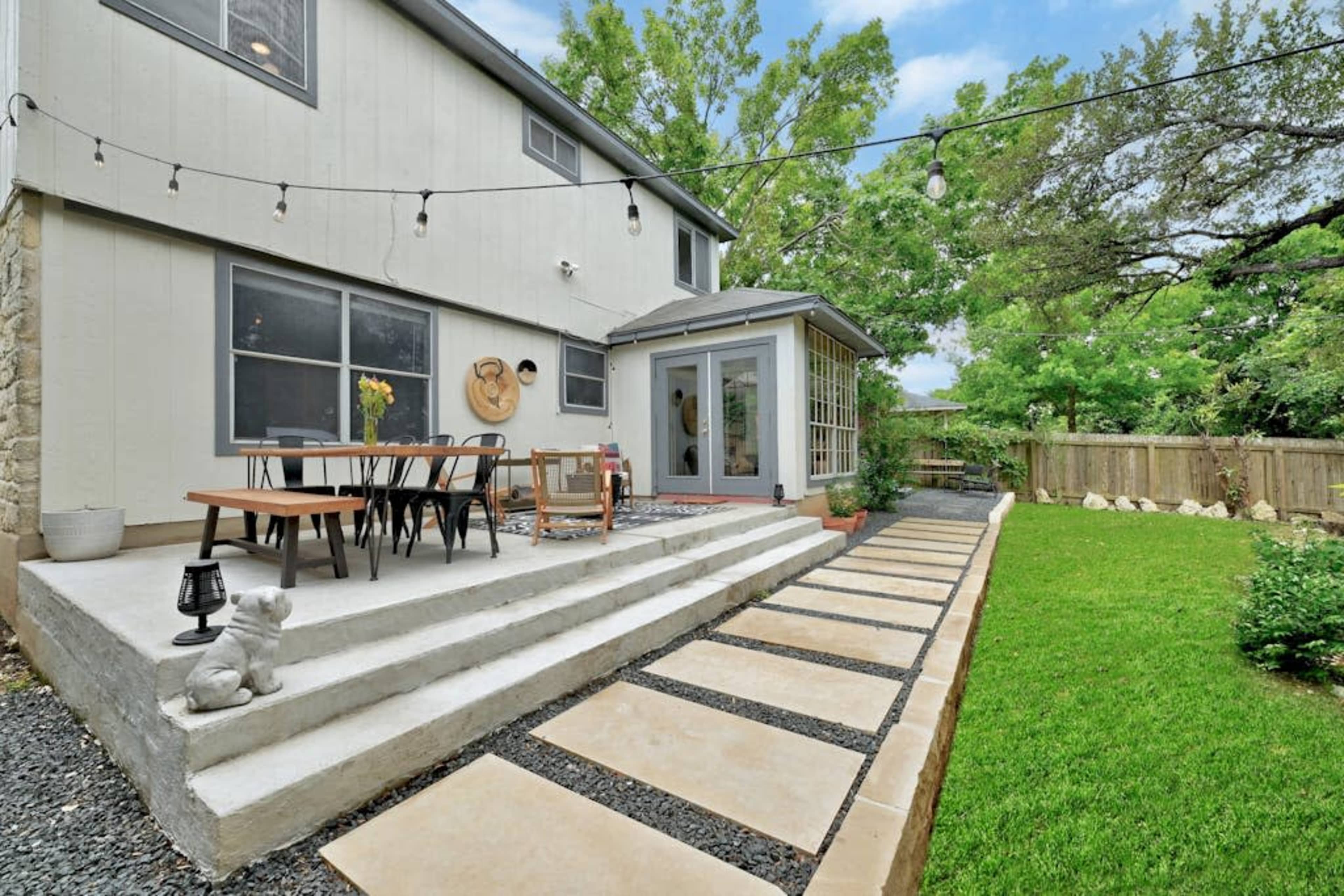 A modern backyard patio features a multi-level stone terrace, wooden dining table, and greenery bordered by a paved walkway.