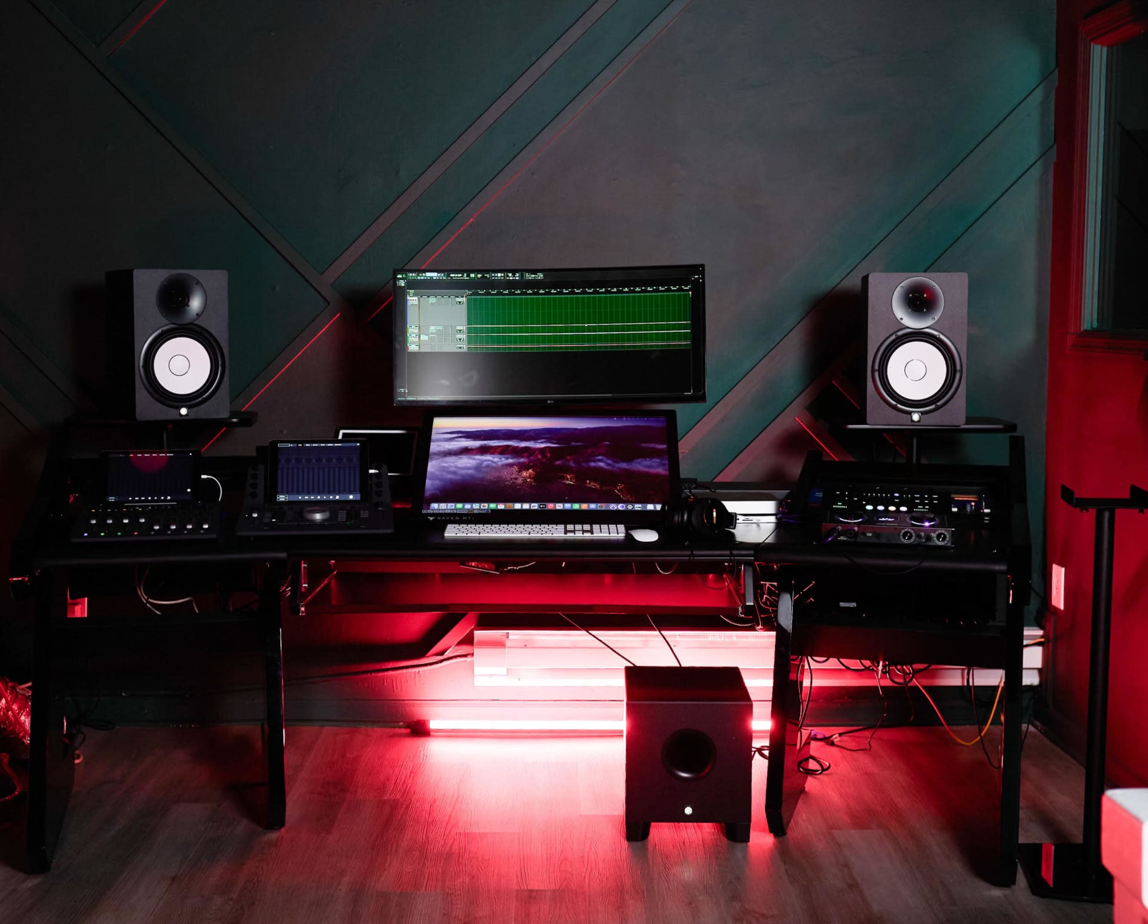 The image shows a music production studio with a large monitor, audio speakers, a digital audio workstation, and various sound equipment on a dark-colored desk.