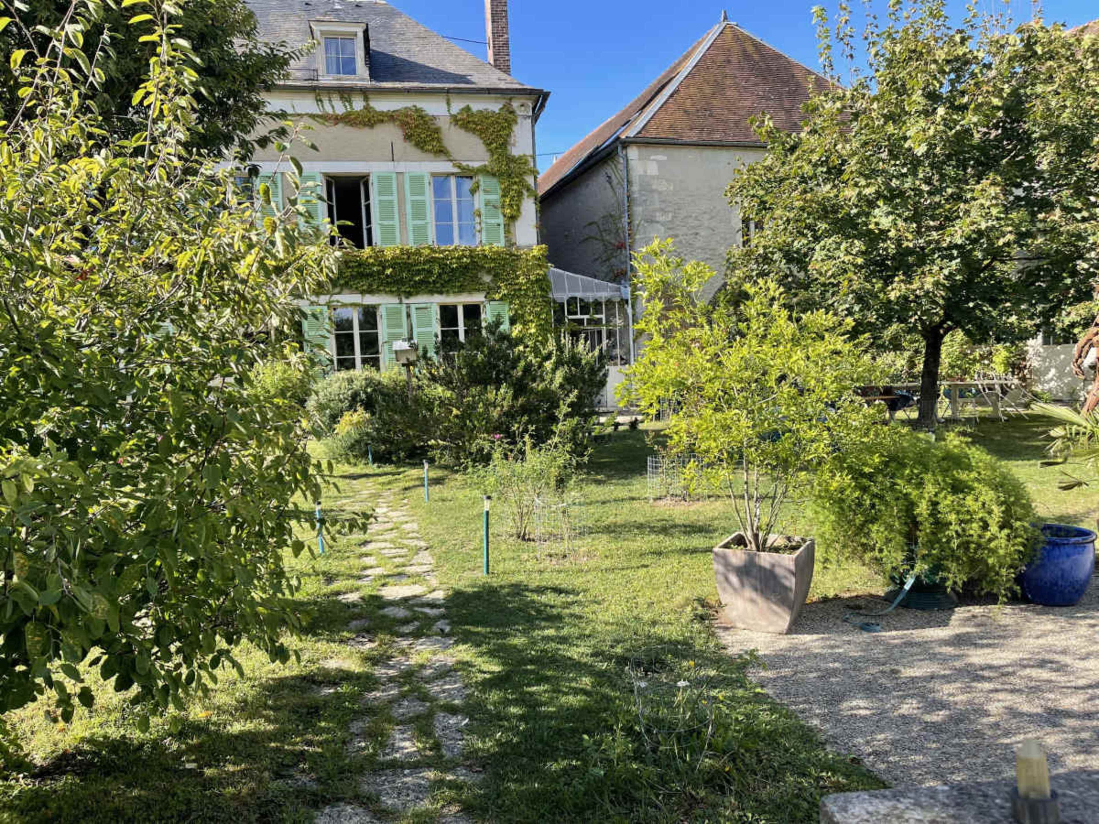 The image shows a house with green shutters, surrounded by a garden featuring various plants and trees under a clear blue sky.