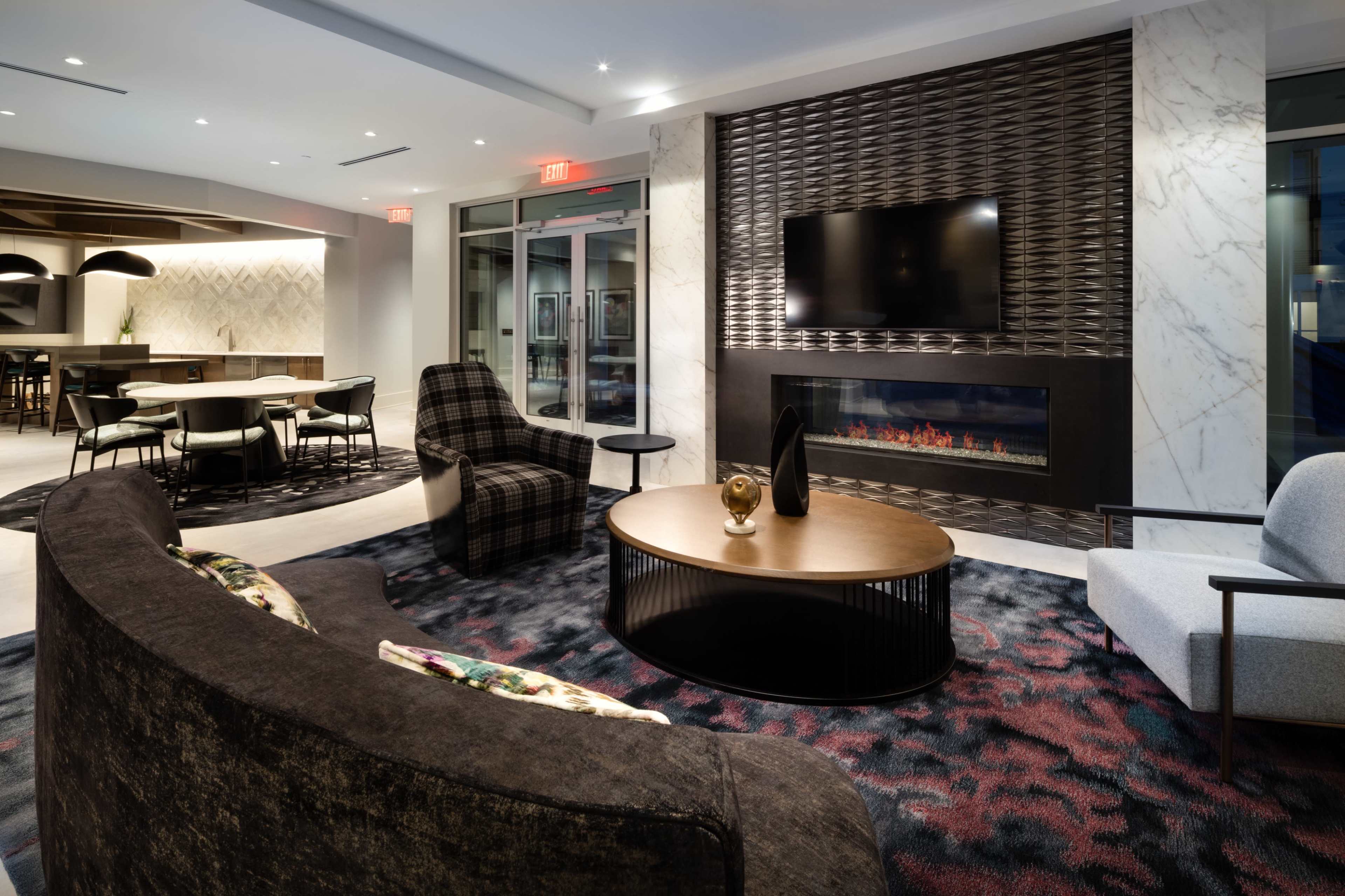 A modern lounge area featuring a curved sofa, a coffee table, a fireplace, and a television mounted on a textured wall, with dining tables and chairs in the background.