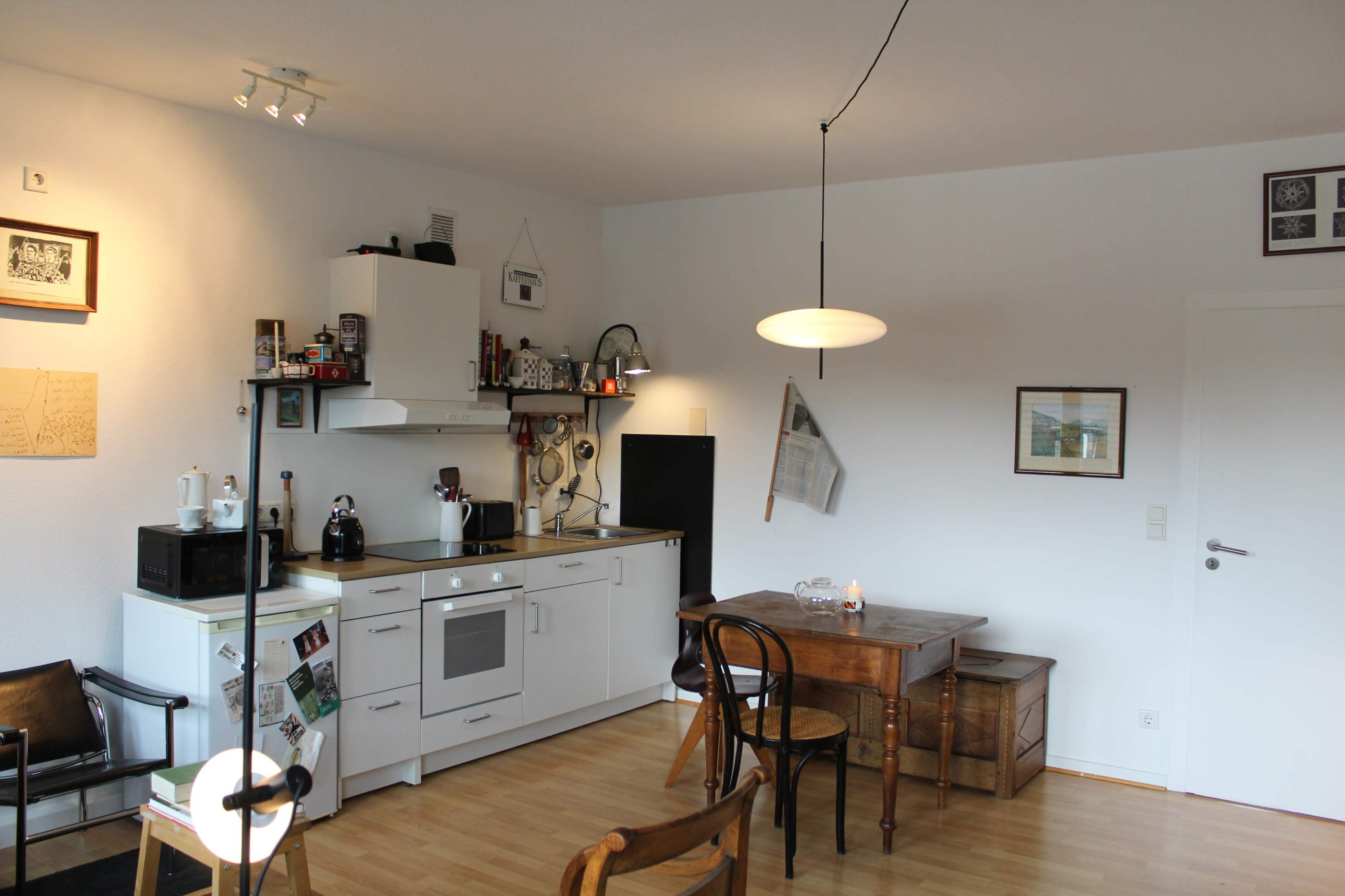 The image shows a compact kitchen area with white cabinets, a small dining table, and a wooden chair, all set in a bright, minimalist room.
