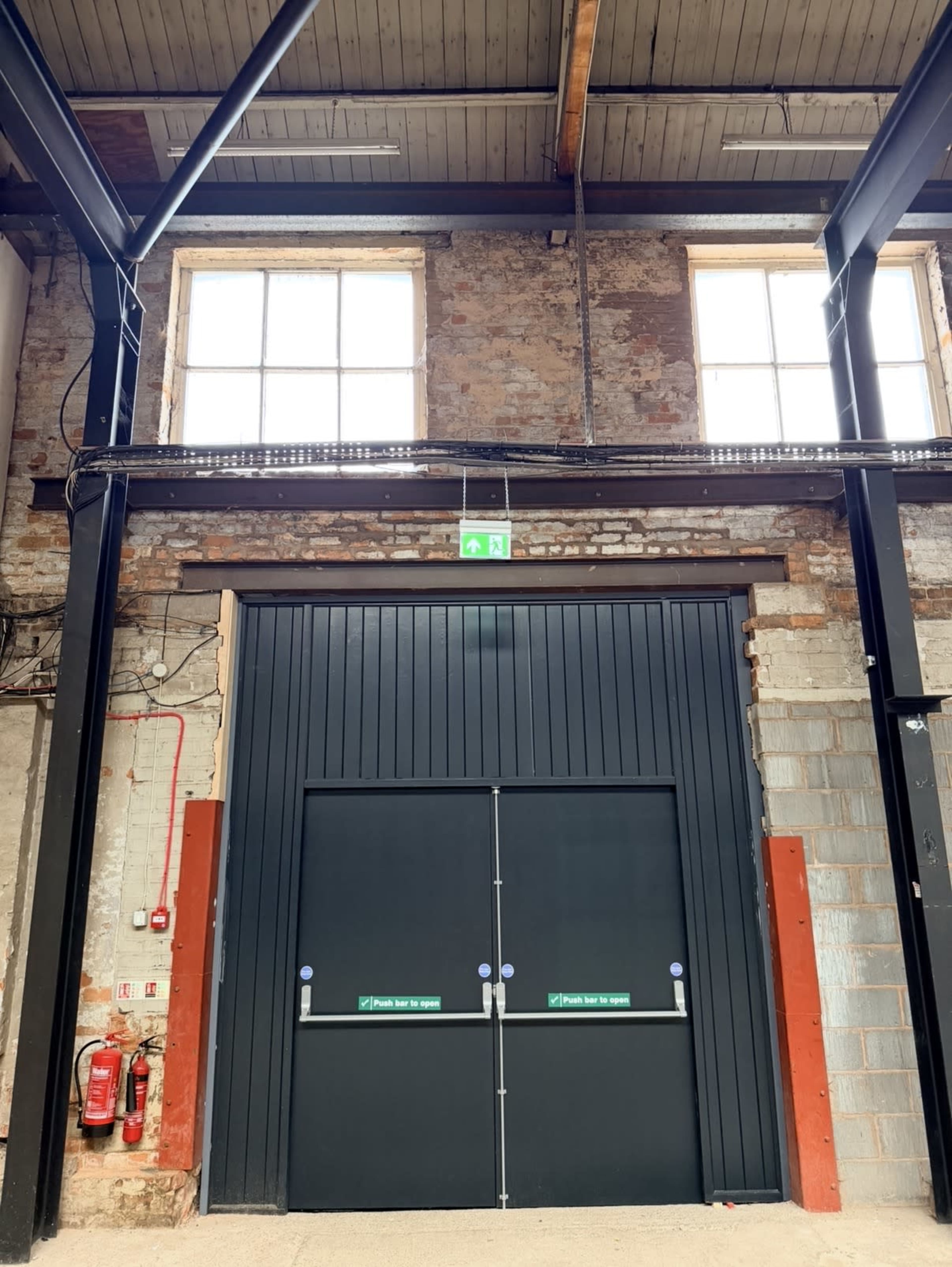 The image shows a large industrial door set against a brick wall with exposed beams and fire safety equipment visible nearby.