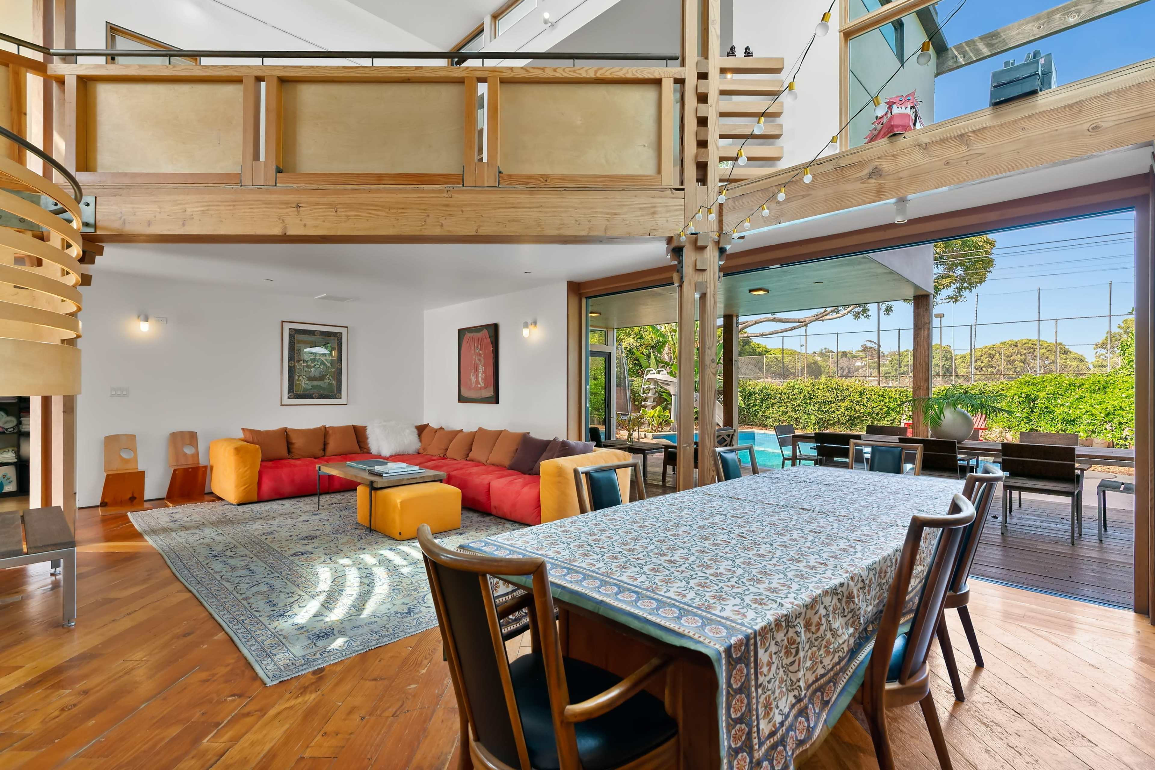 The image shows a spacious, modern interior with a dining area featuring a patterned tablecloth, a cozy living space with a red sofa, and large windows that provide natural light and a view of greenery outside.