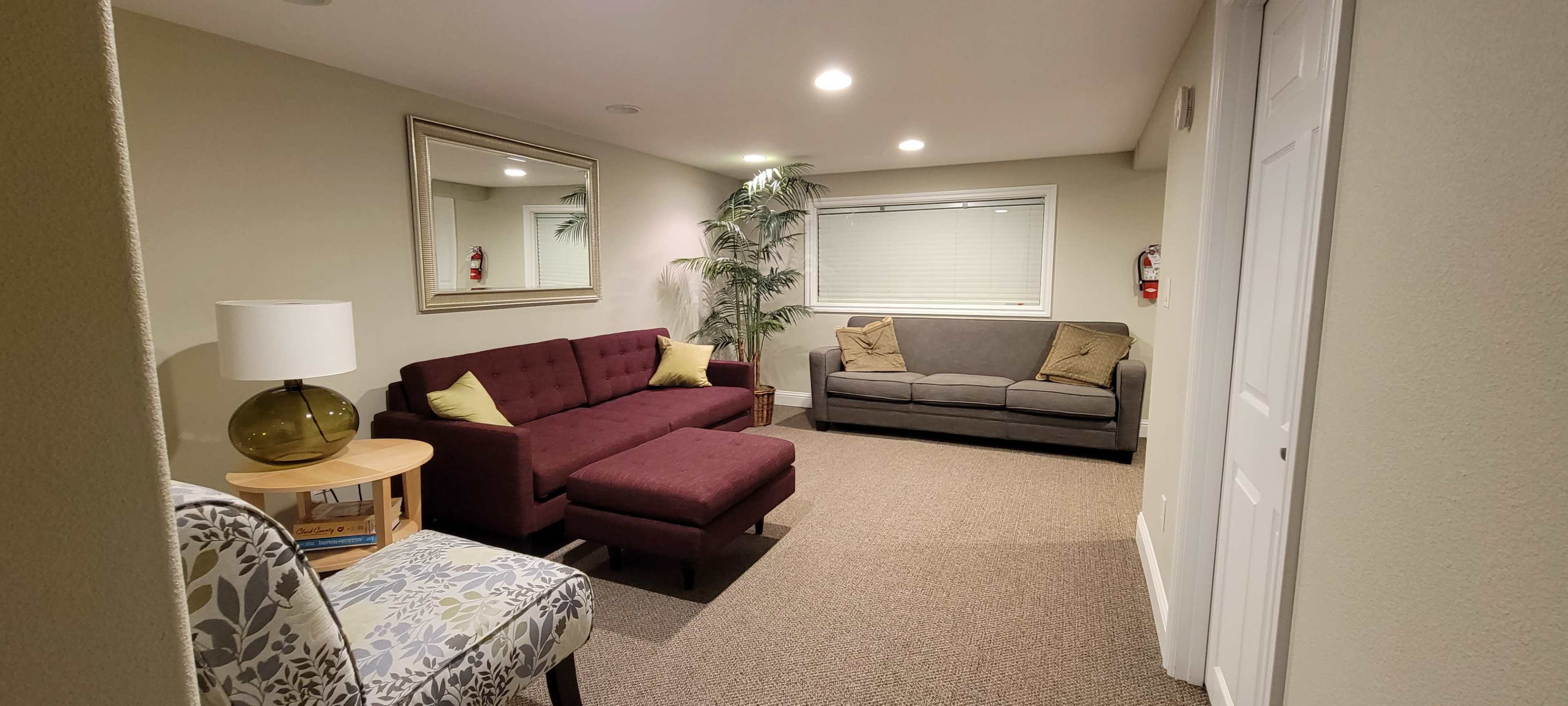 A cozy living room with a maroon sectional sofa, a gray couch, a small table with a lamp, and a decorative chair against beige walls.