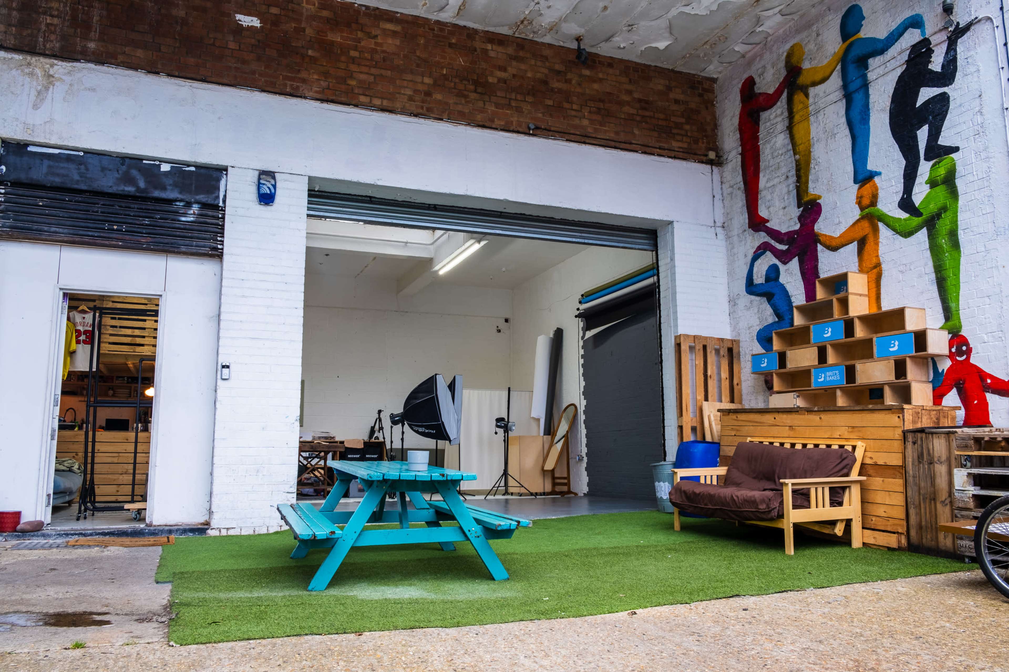 An outdoor area of a warehouse featuring a turquoise picnic table, a brown couch, and colorful murals of people on the wall.