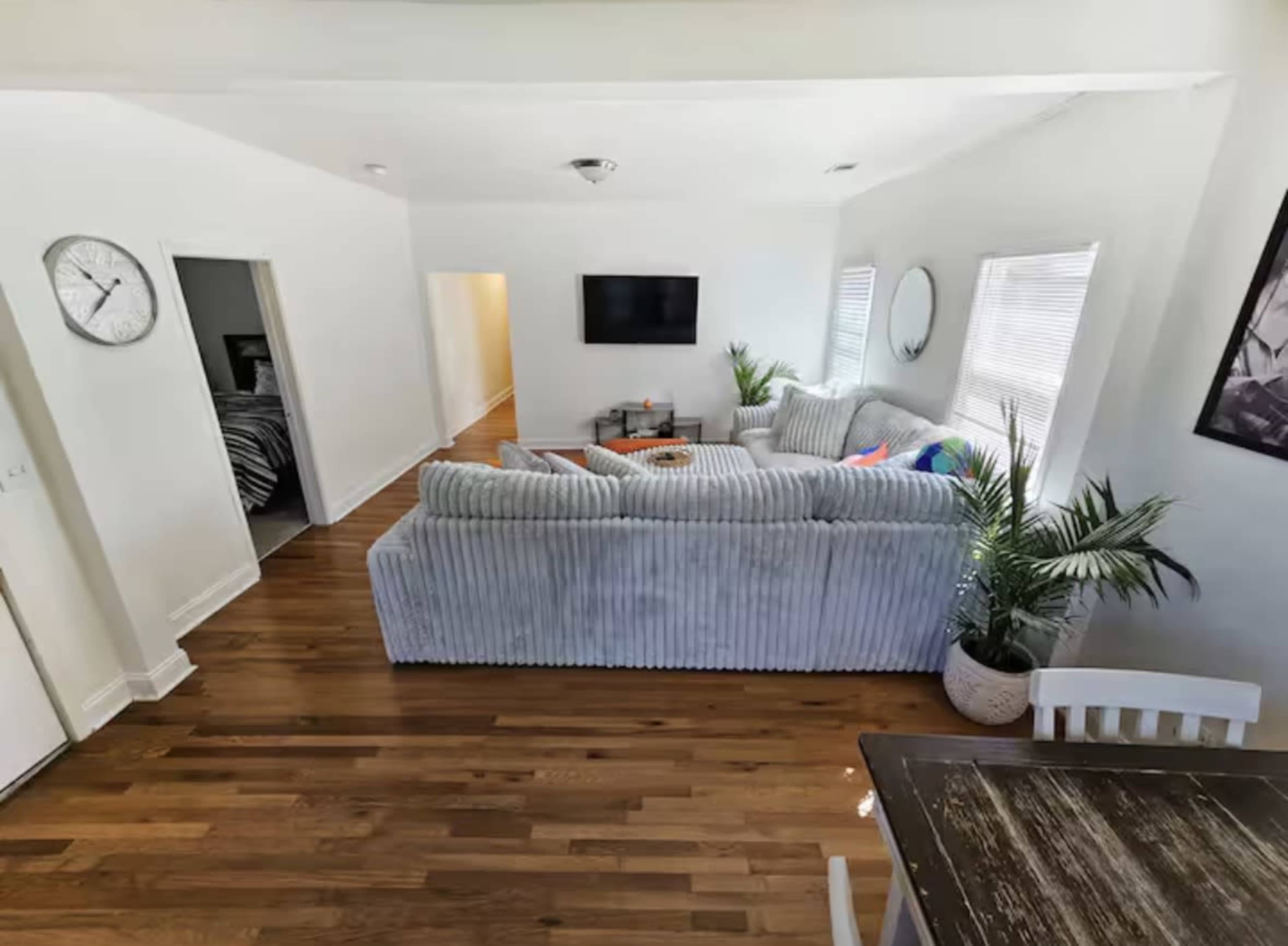 The image shows a light-filled living room featuring a sectional sofa, a coffee table, a television mounted on the wall, and potted plants, with a dining area visible in the foreground.
