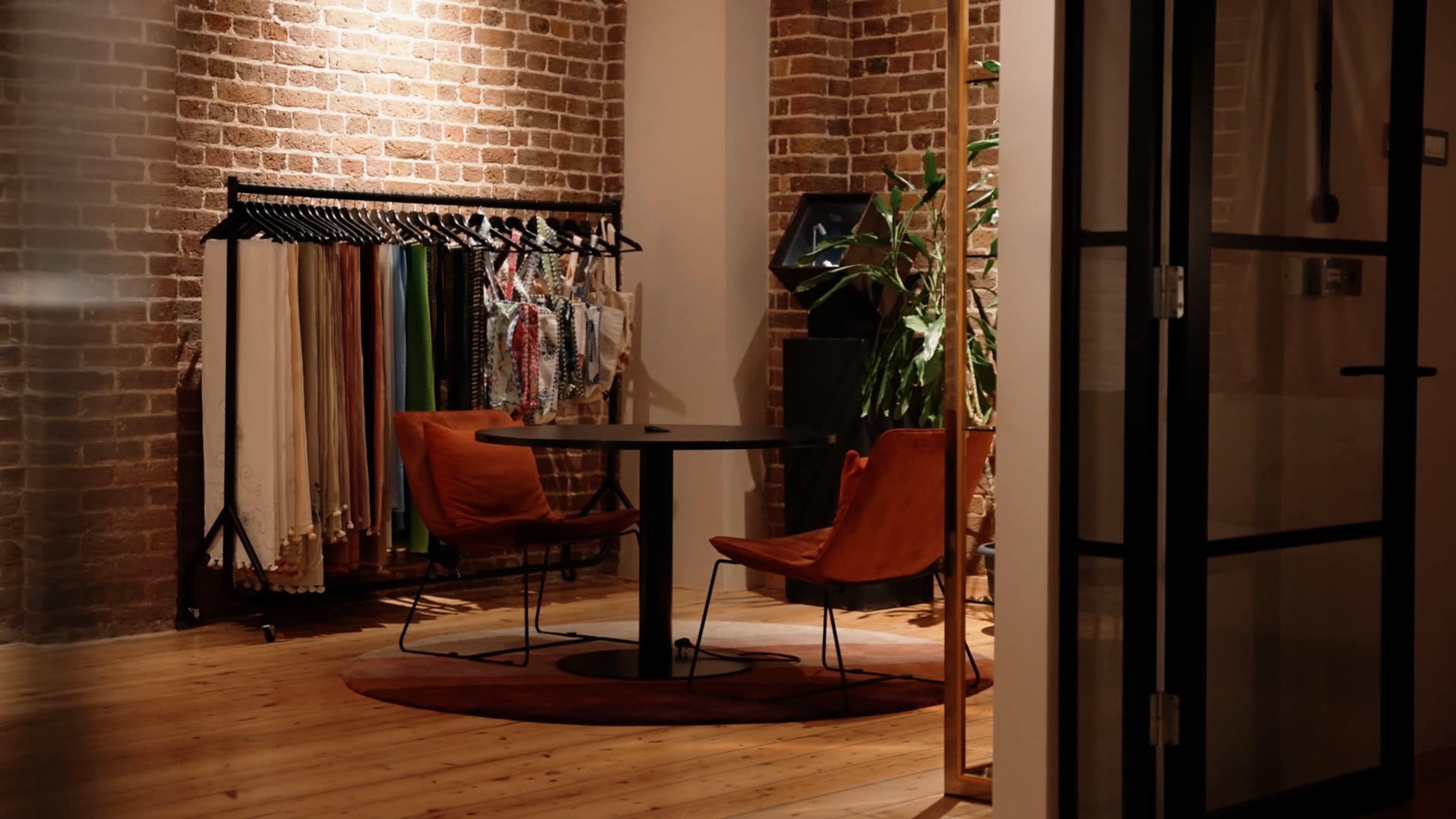 The image shows a cozy room with exposed brick walls, featuring a round table and two chairs, alongside a rack with various textiles.