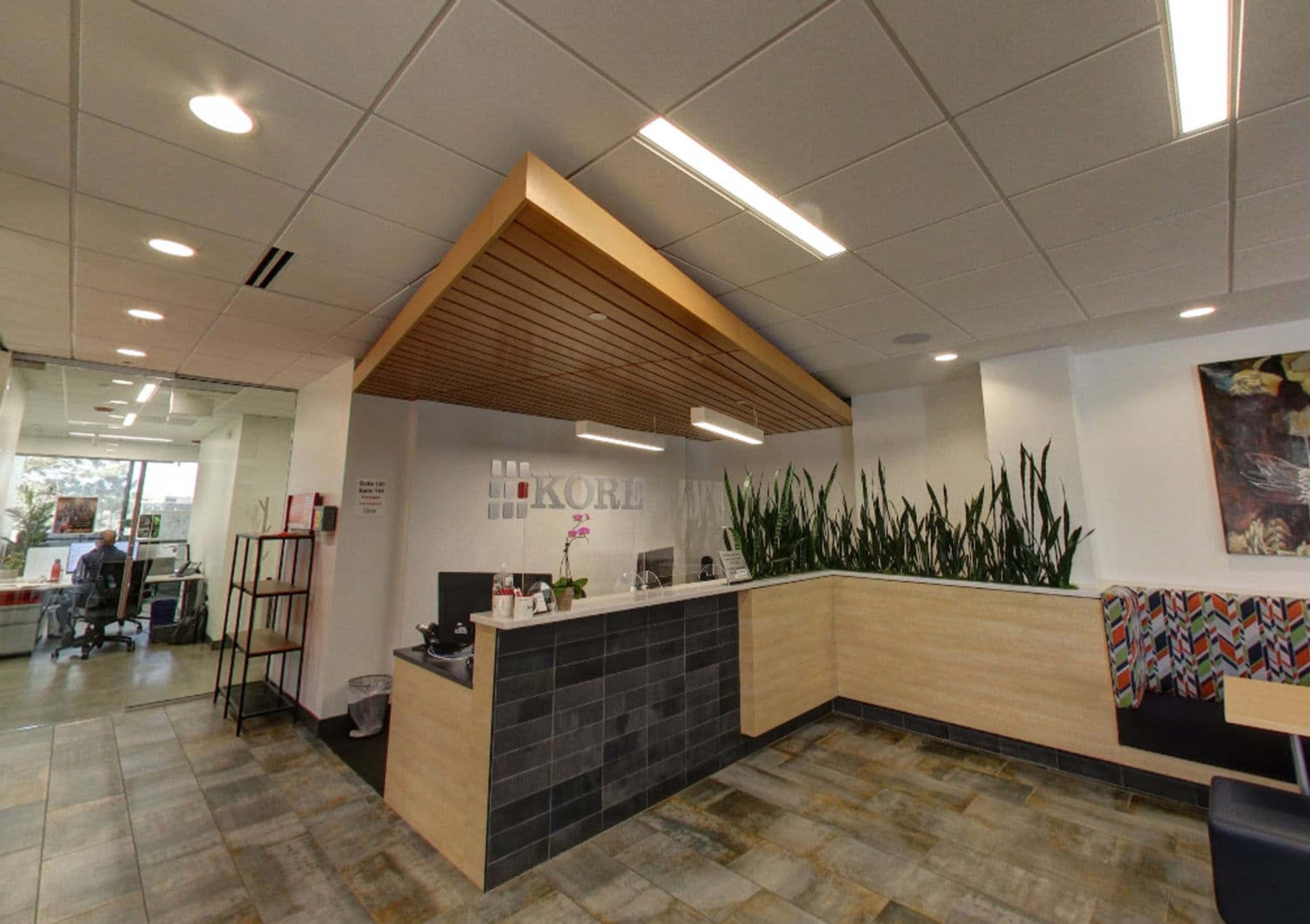 The image shows a modern reception area with a wooden ceiling feature, a sleek front desk, and green plants along the wall.
