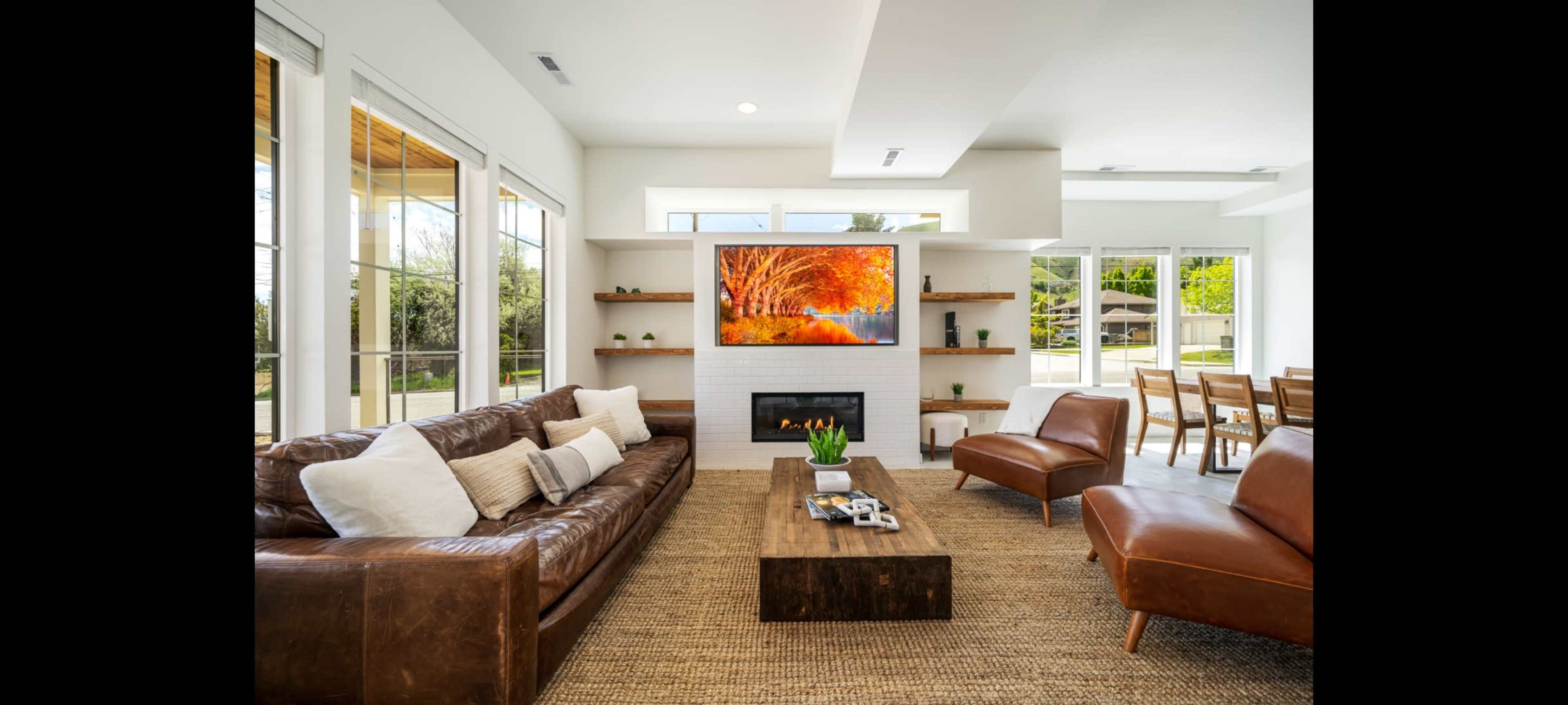 A modern living room features a brown leather sofa, a wooden coffee table, and a large wall-mounted television displaying an autumn scene.