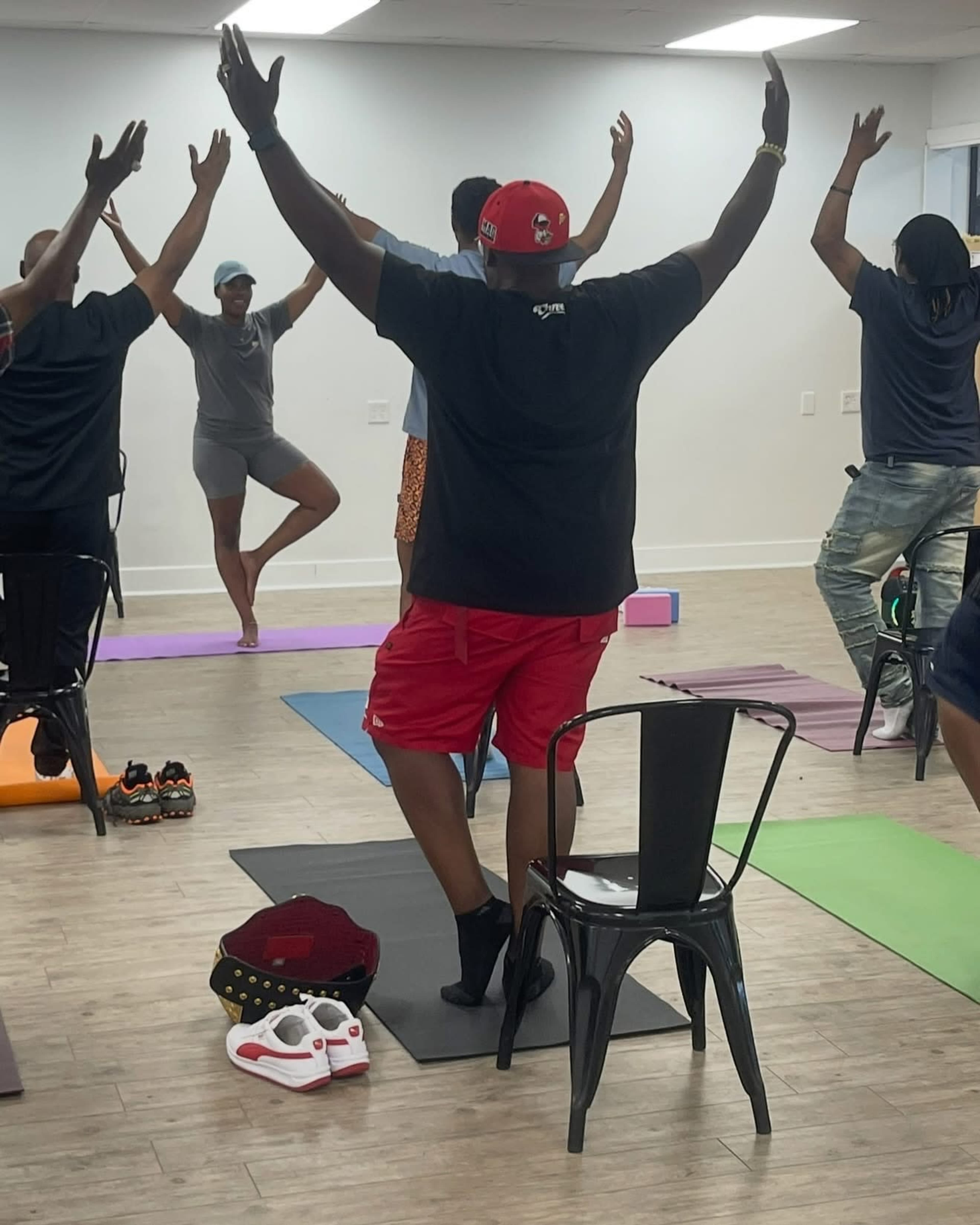 In a brightly lit room, several individuals are participating in a chair yoga class, with one person balancing on one leg while seated on a chair.