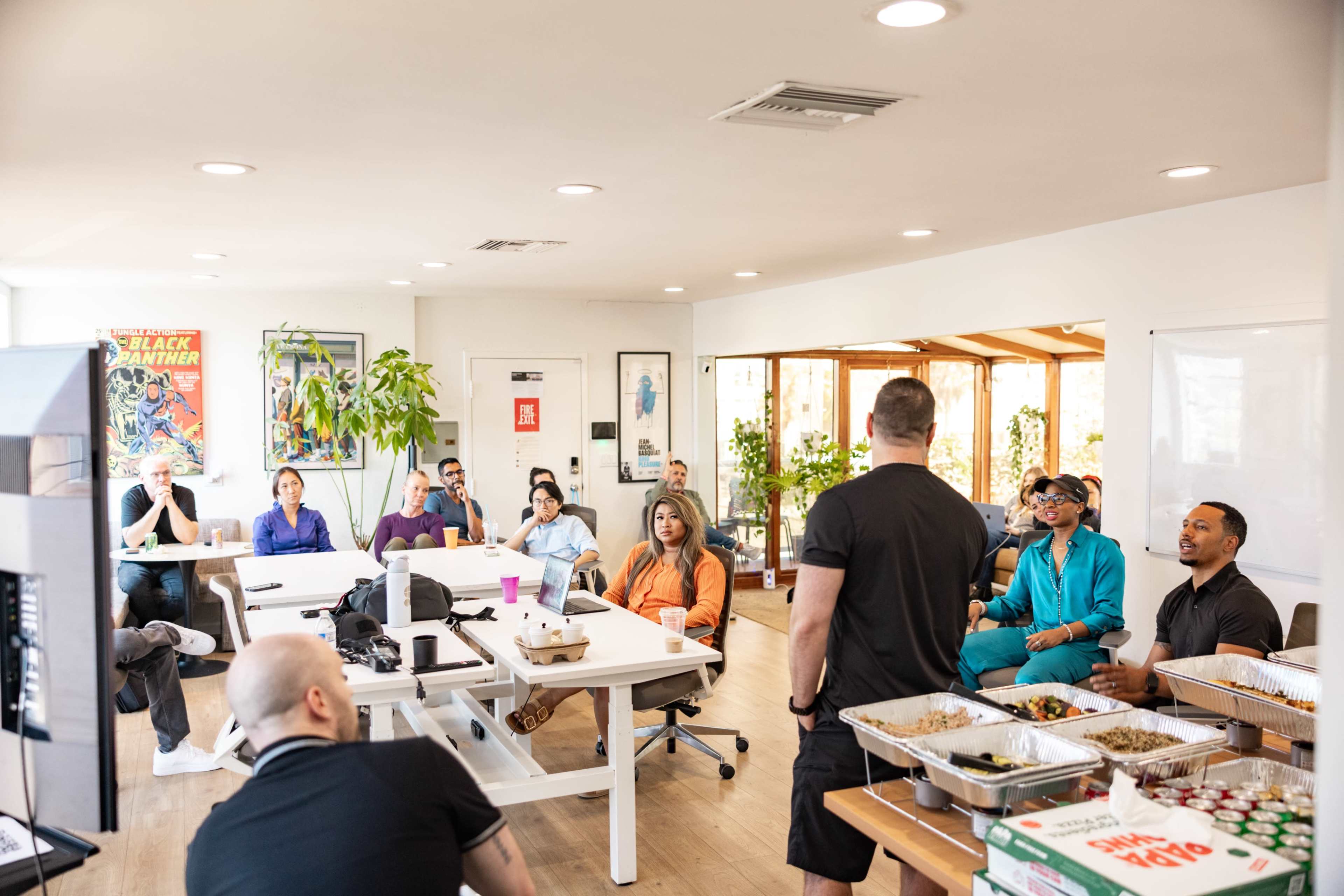 A group of people is seated in an office space during a presentation, with food trays on tables and large windows letting in natural light.