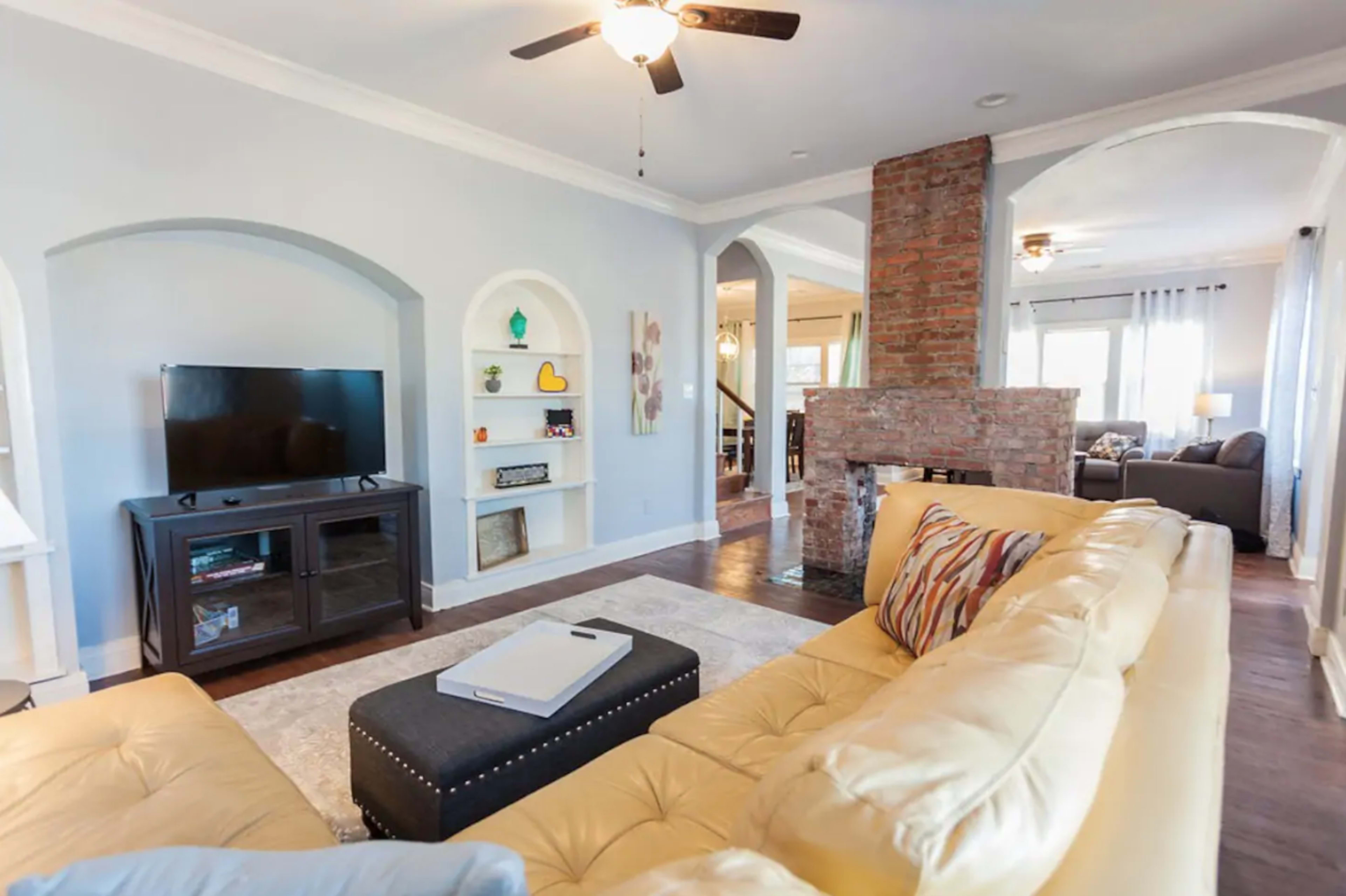 The image shows a modern living room featuring a yellow sectional sofa, a mounted television, and a brick fireplace, with open archways leading to other areas of the home.