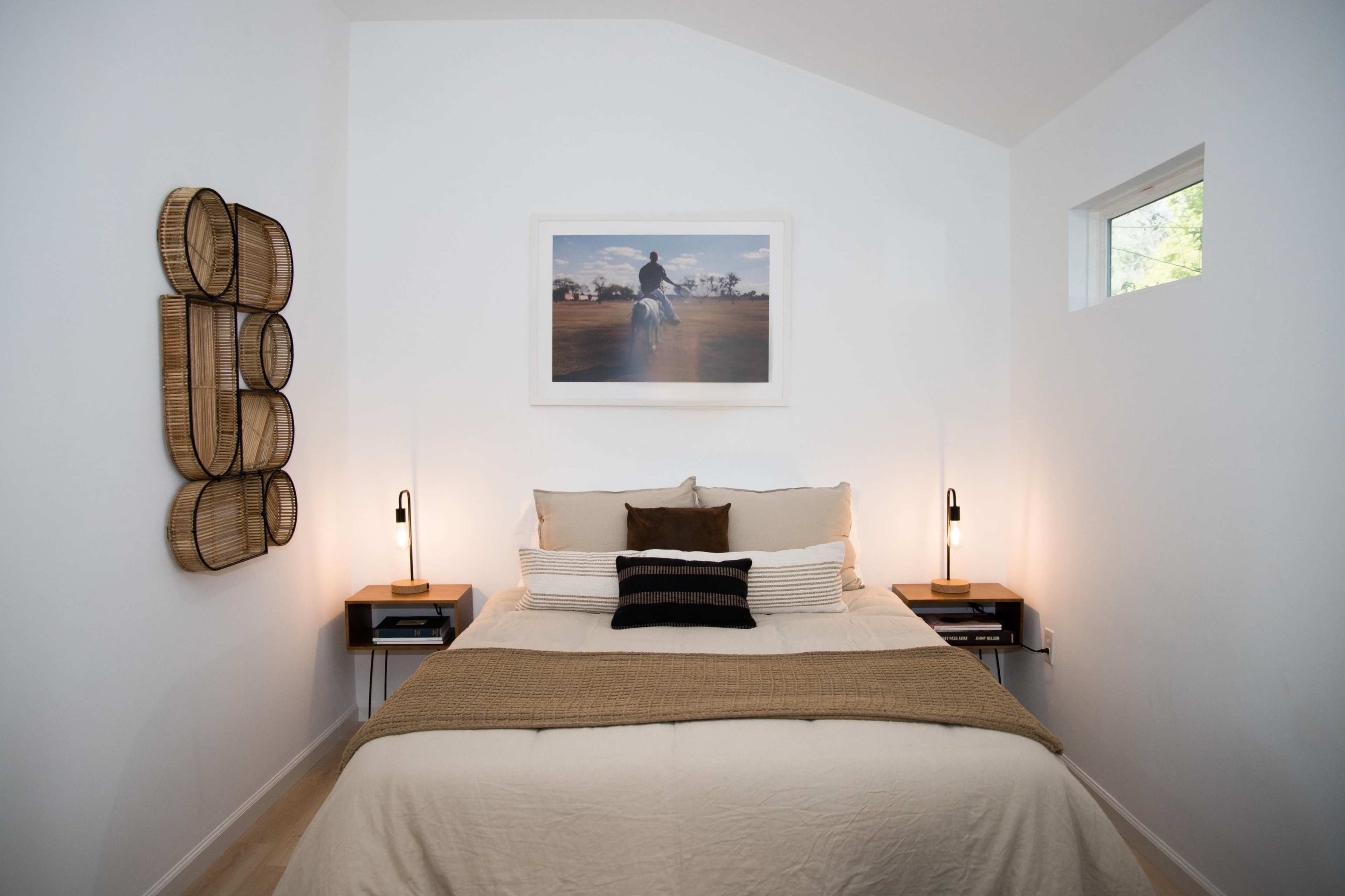 The image shows a minimalist bedroom with a bed, decorative wall art, two bedside lamps, and a woven wall hanging.