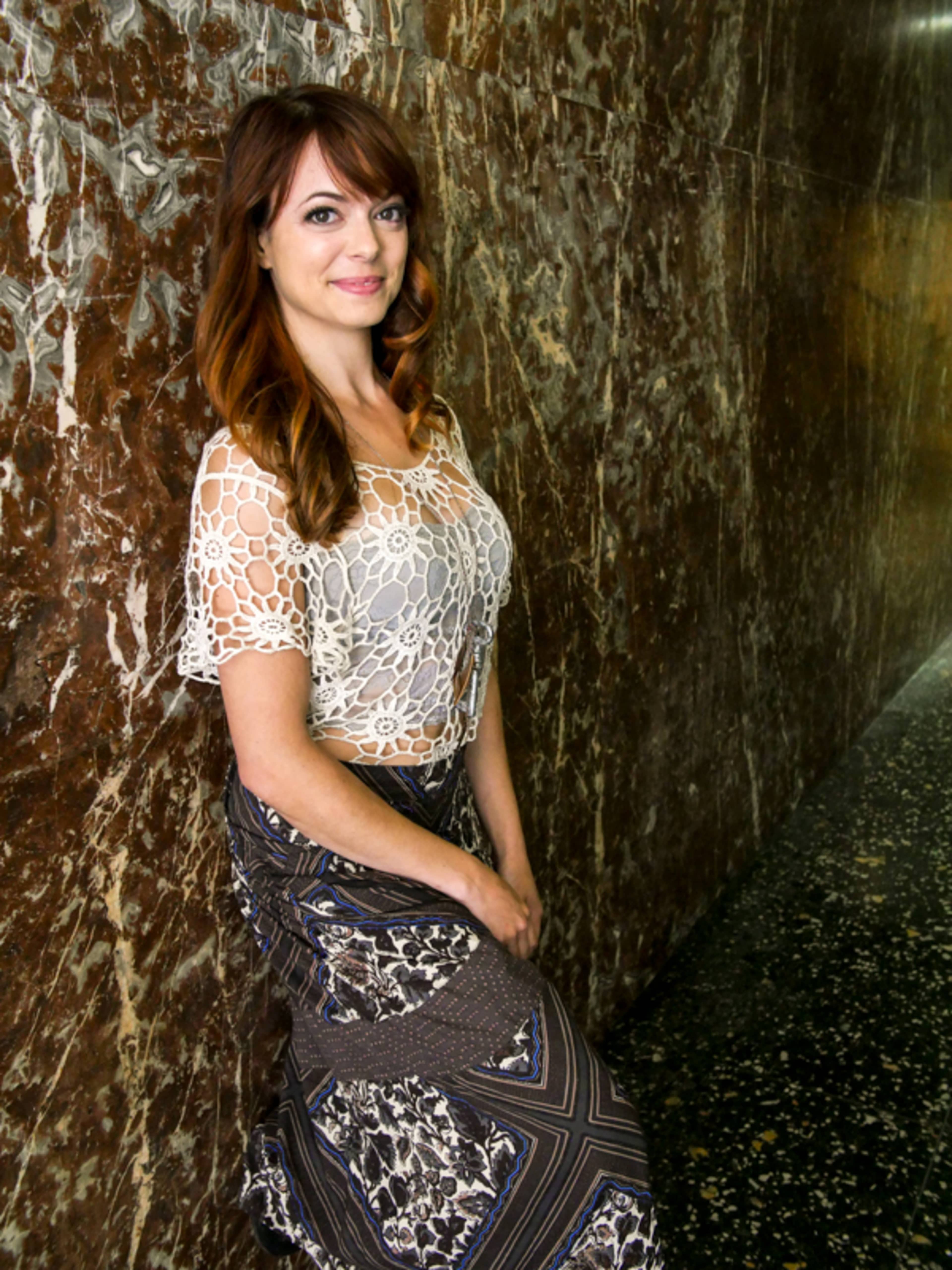 A woman stands against a textured stone wall, dressed in a lace top and a patterned skirt.