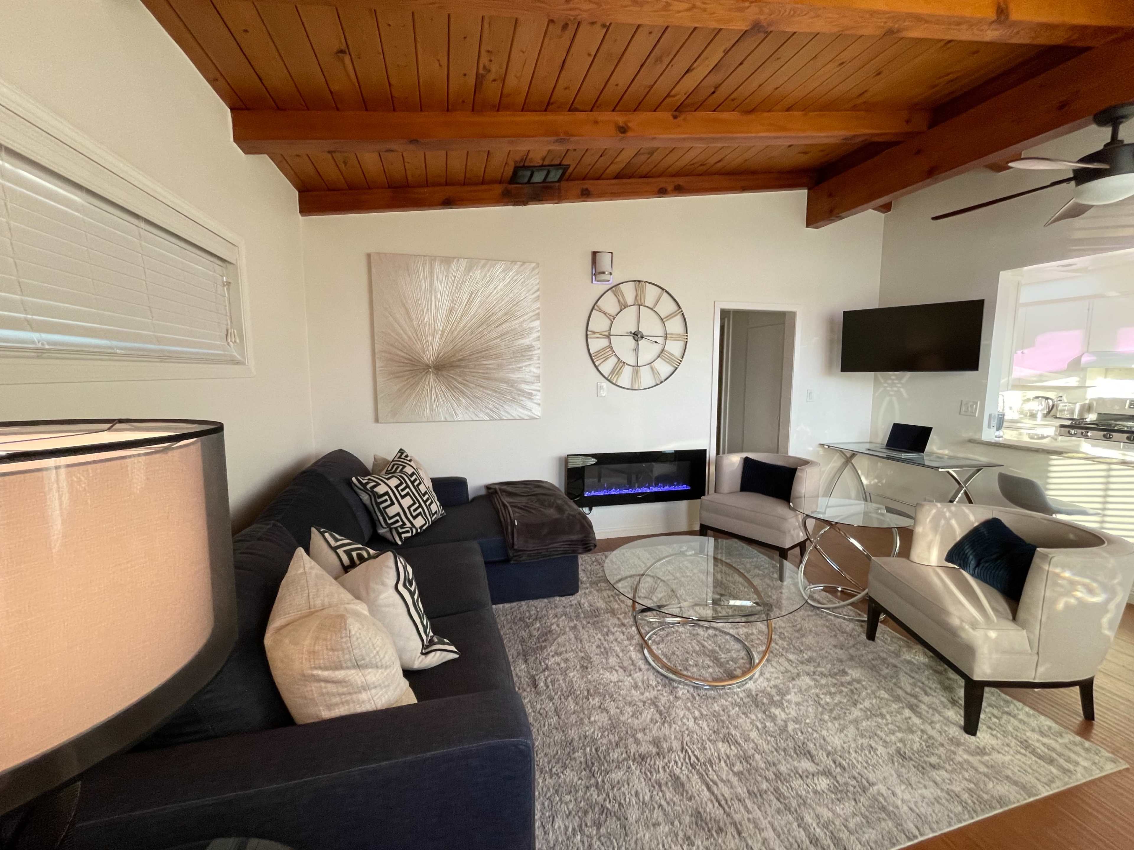 The living room features a dark sofa arrangement, a glass coffee table, an electric fireplace, and a modern art piece on the wall under wooden ceiling beams.