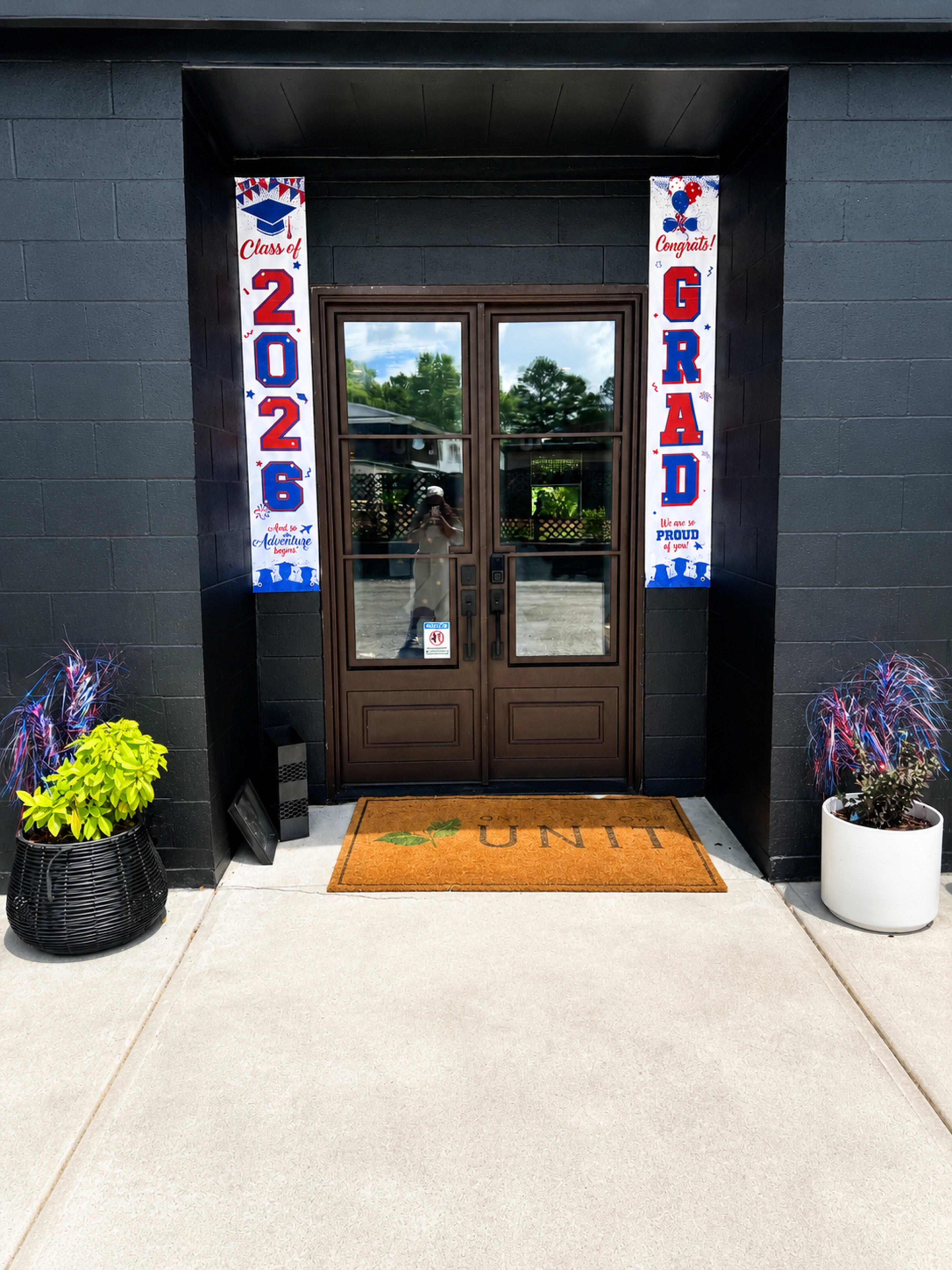 The entrance features a pair of wooden double doors flanked by graduation banners and potted plants on either side.