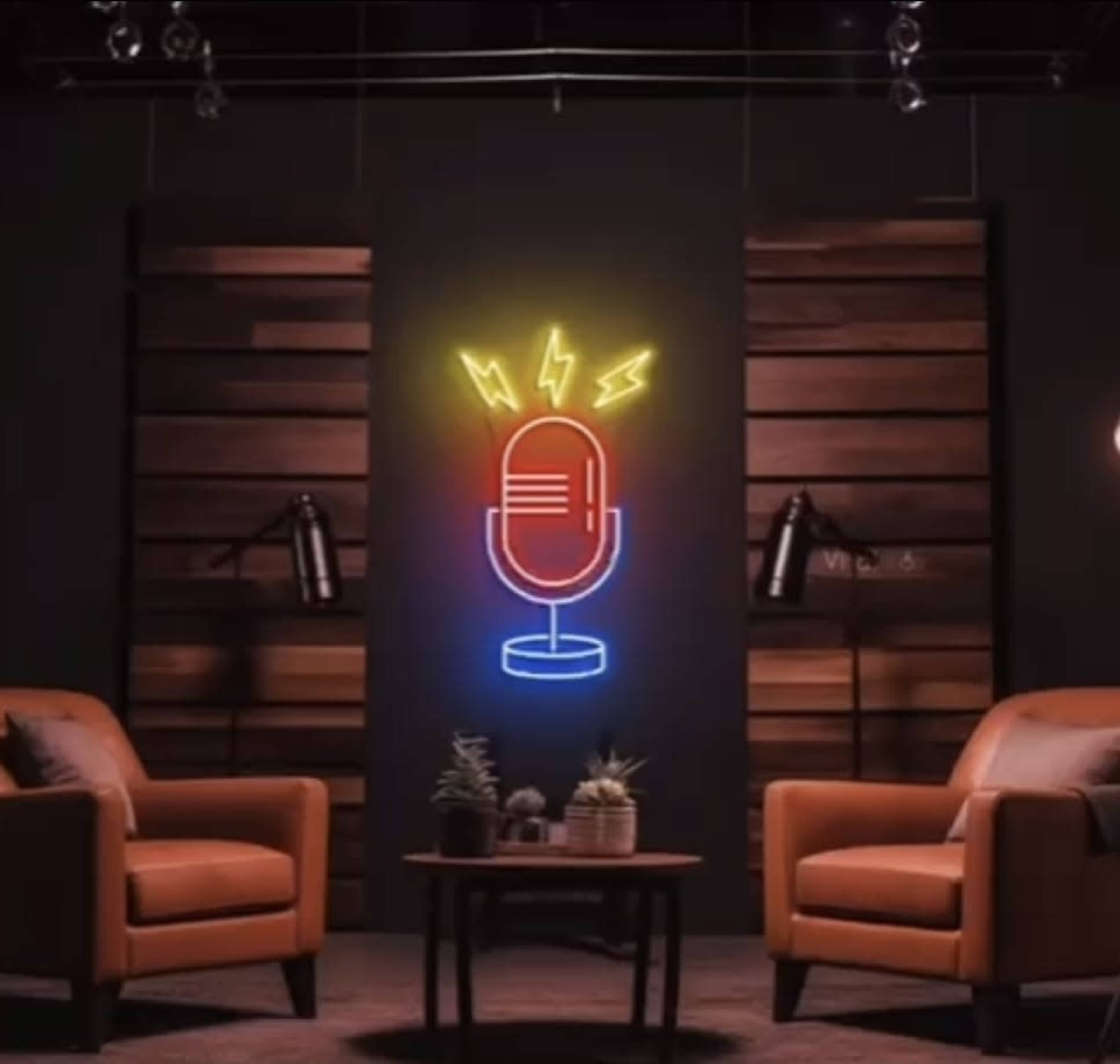 The image features a podcast studio setup with two brown chairs facing a small table, a neon microphone sign on the wall, and pendant lights overhead.