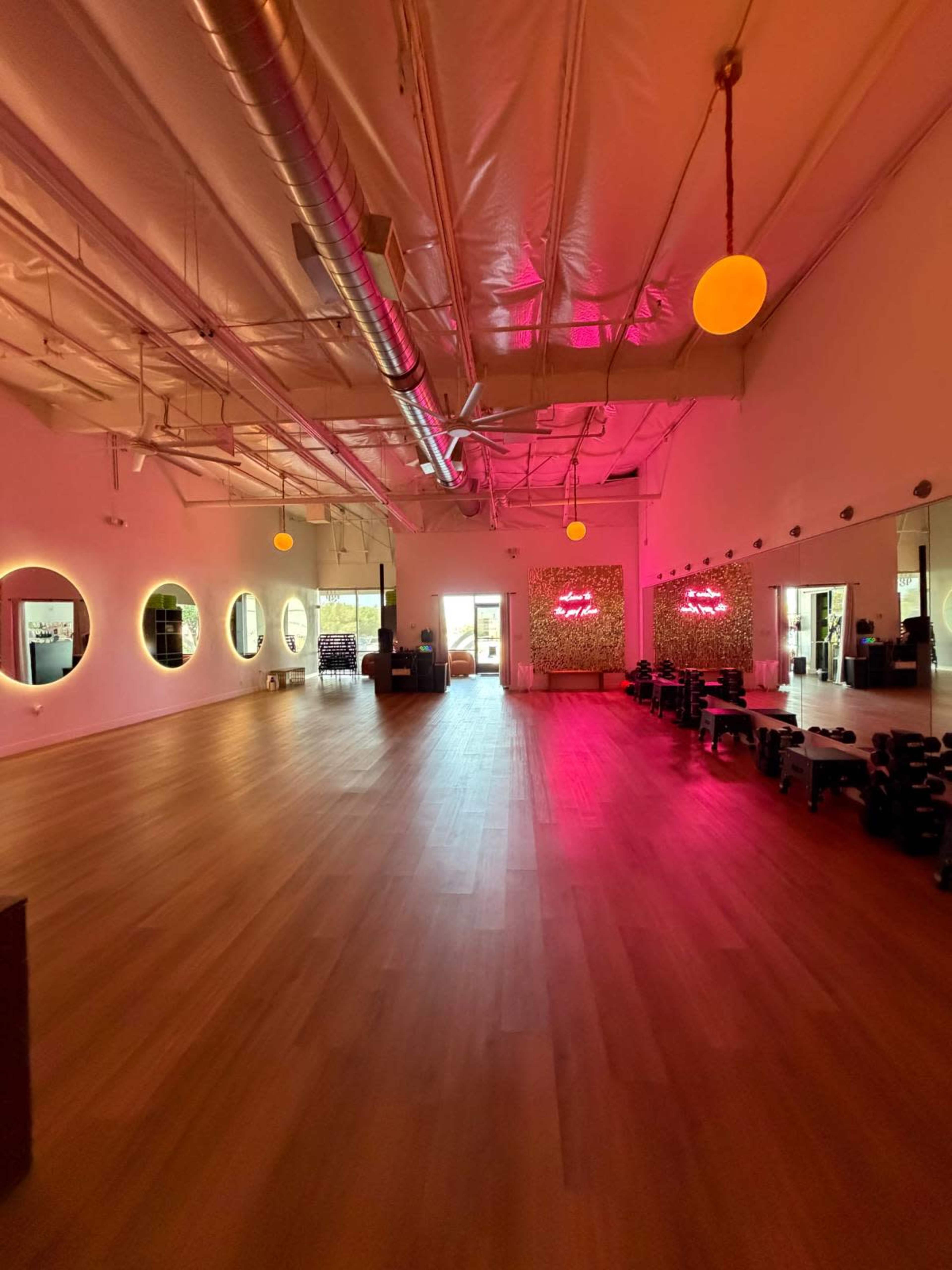 The image shows a spacious fitness studio with wooden flooring, large mirrors, and neon lighting accents.