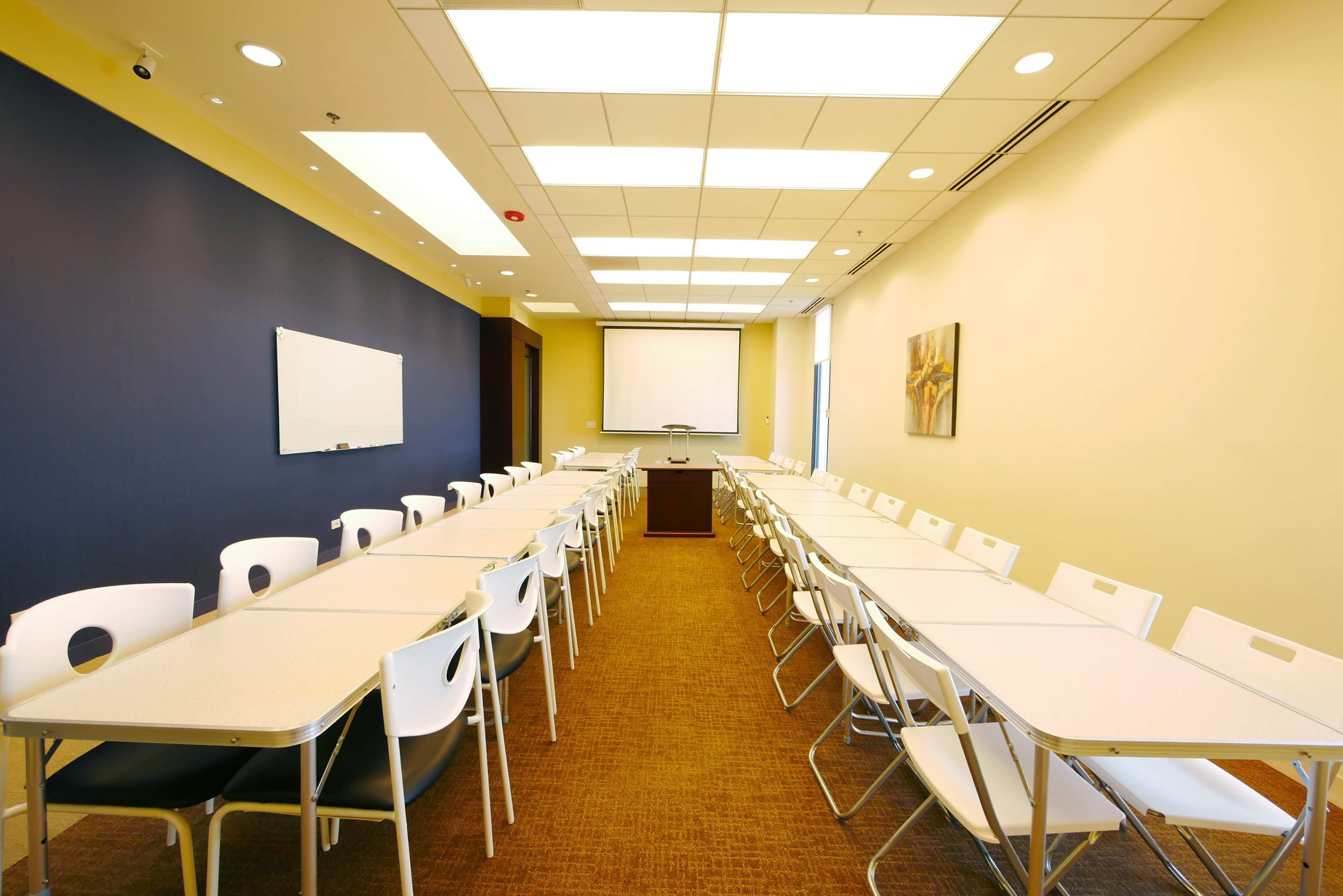 A classroom setup features several rows of tables and chairs facing a front presentation area with a screen and whiteboard.