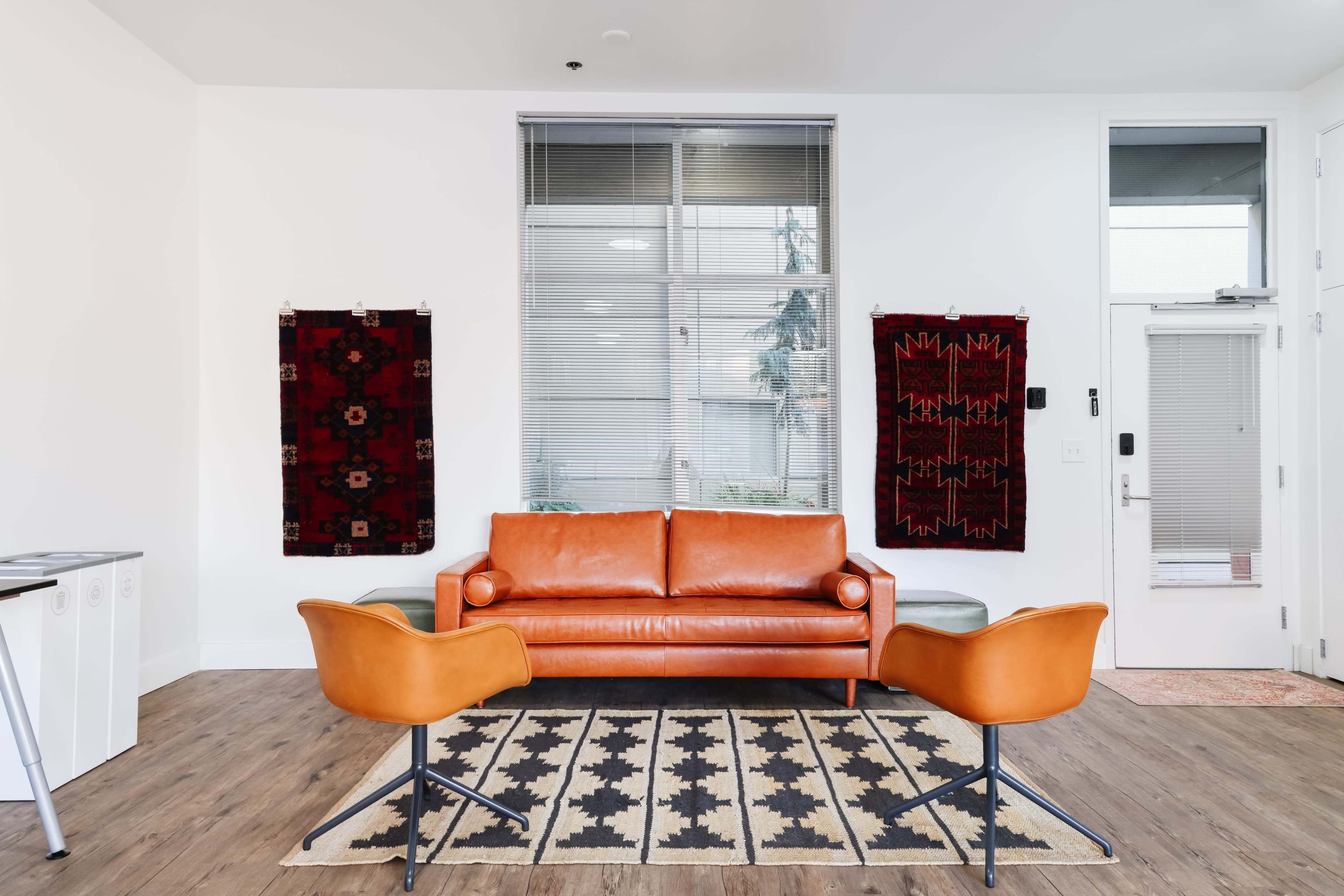 The scene features a modern living room with an orange leather sofa, two contemporary chairs, and patterned rugs hanging on the walls.