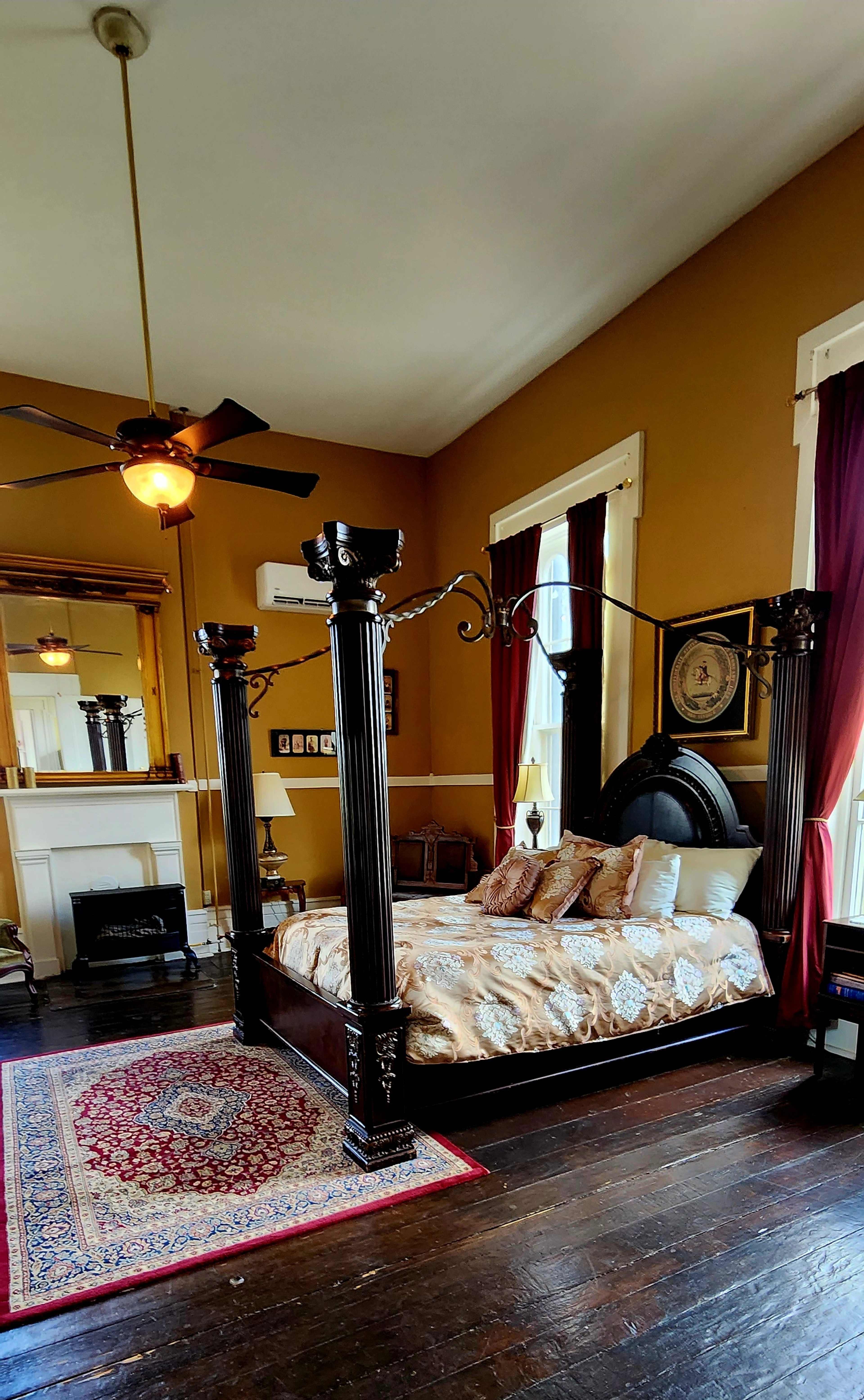The room features a four-poster bed, a decorative mirror, and a patterned area rug against a gold-painted wall.